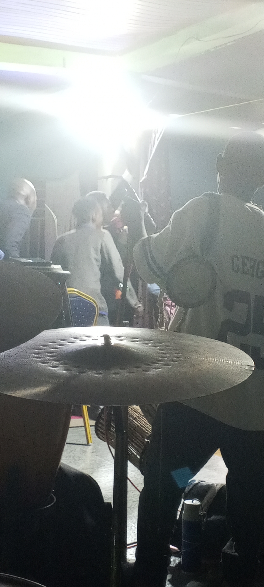 The image captures a dimly lit indoor scene featuring a drum set, musicians, and onlookers. A cymbal dominates the foreground, positioned on a stand with a woven drum visible behind it. In the middle ground, figures are present, including a person wearing a light-colored shirt with a "GERG" inscription and the number "25," and other individuals, some with obscured faces. A blue chair is partially visible. The background is blurred with bright lights creating an indistinct atmosphere.