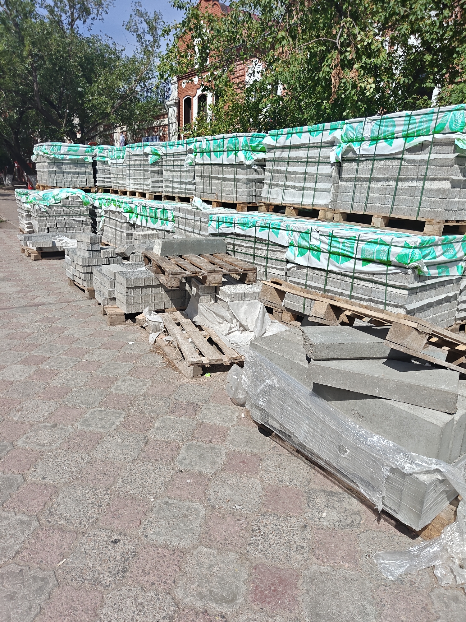A paved street in the city of Petropavl, Kazakhstan is shown, lined with stacks of grey concrete paving stones. The stones are mostly organized on wooden pallets, some wrapped in green and white plastic sheeting. The paving stones are of various sizes and shapes, with some individual stones and some bundles visible. In the background, a red brick building with arched windows and trees with green foliage are visible, suggesting a daytime scene under a clear sky. The street itself is paved with interlocking concrete pavers in a reddish-pink and grey pattern.