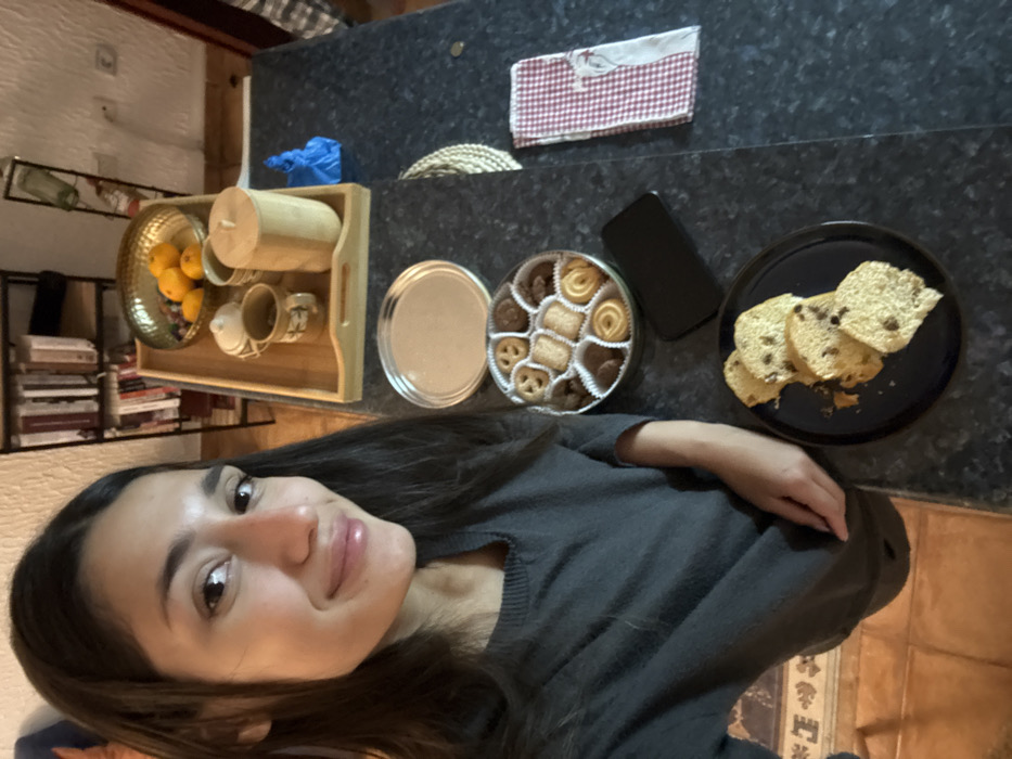 A person is at a dark granite countertop. On the counter is a wooden tray containing oranges, blueberries, a tea set, a tin of assorted cookies, a black cell phone, a black plate of raisin bread slices, and a red and white checkered cloth. A bookshelf with books is visible in the background.