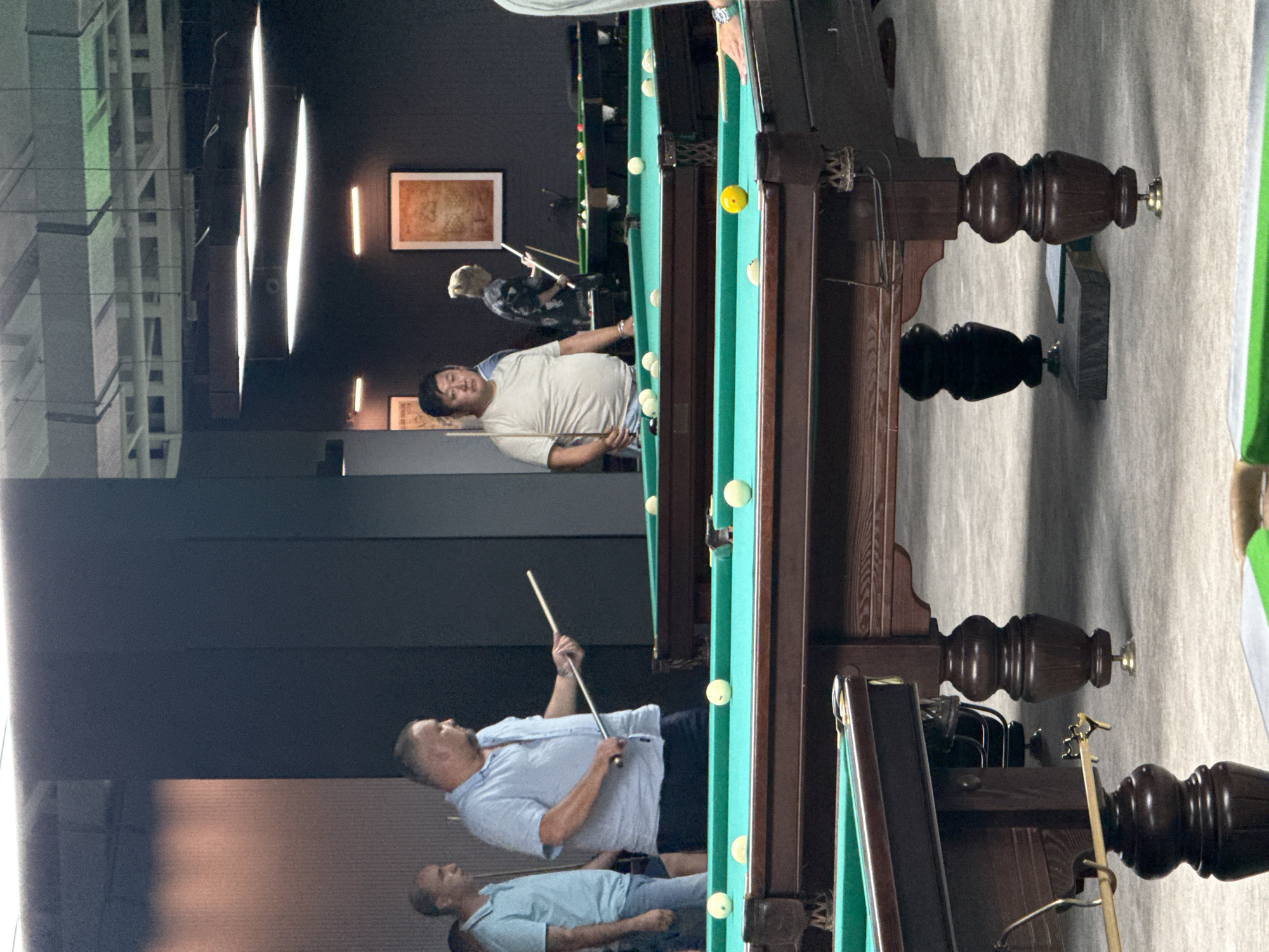 The image depicts an indoor billiards hall in the Balaklavsky District of Ukraine. Several people are present, engaged in or observing a game of billiards. In the foreground, a pool table with green felt is visible, with several white balls scattered across its surface. A yellow ball is also present. A person's hand is visible on the right side, suggesting they are about to take a shot or have just finished one. In the midground, another pool table is set up, with a man in a light blue shirt holding a cue stick, seemingly in mid-game. Another man in a light blue polo shirt stands to his left, observing. Further back, a man in a beige shirt stands near a pool table, also with a cue stick. The environment appears to be a modern establishment, with dark walls and ceiling lights that illuminate the playing area. The overall impression is one of a social gathering centered around the game of billiards.
