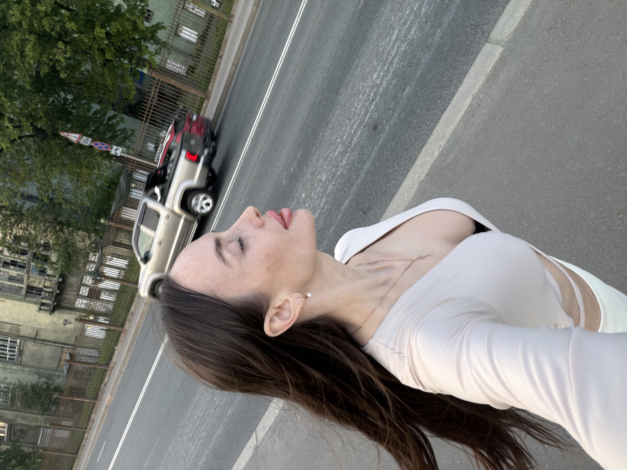 An outdoor, eye-level selfie captures a young adult female on a city street in Saint Petersburg, Russia. The woman, positioned prominently on the left, has long, flowing dark brown hair and light skin with visible freckles on her forehead and cheek. Her eyes are closed or averted to her right (viewer's left), and her lips are pink. She wears a light, possibly pale pink or nude, long-sleeved top with a v-neckline revealing a black bra, and cut-out details on the sides. A thin gold-colored chain with a small cross pendant hangs around her neck, and a small gold-colored hoop earring with a white stone is in her right ear. White clothing is visible at the bottom of the frame.
To her right, a two-lane asphalt road with double solid white lines separates her from the opposite side of the street. A concrete curb marks the edge of the pedestrian area. A silver/light gold Volkswagen Amarok pickup truck, with a red vehicle partially visible in or behind its bed, travels from left to right on the road, appearing slightly blurred due to motion.
Beyond the road, a metal fence runs parallel to the street, behind which stand large, multi-story, green-hued historical buildings. These structures exhibit architectural details like multiple windows, some appearing old or boarded, and a decorative balcony on the building to the right. Dense green trees are interspersed among and behind the buildings. A utility pole on the far side of the street displays three vertical traffic signs: a red triangular warning sign with an exclamation mark and "300m" text, a blue circular "No Stopping" sign with a red diagonal line, and a blue square sign with a white arrow indicating direction. The lighting suggests late afternoon or early evening.