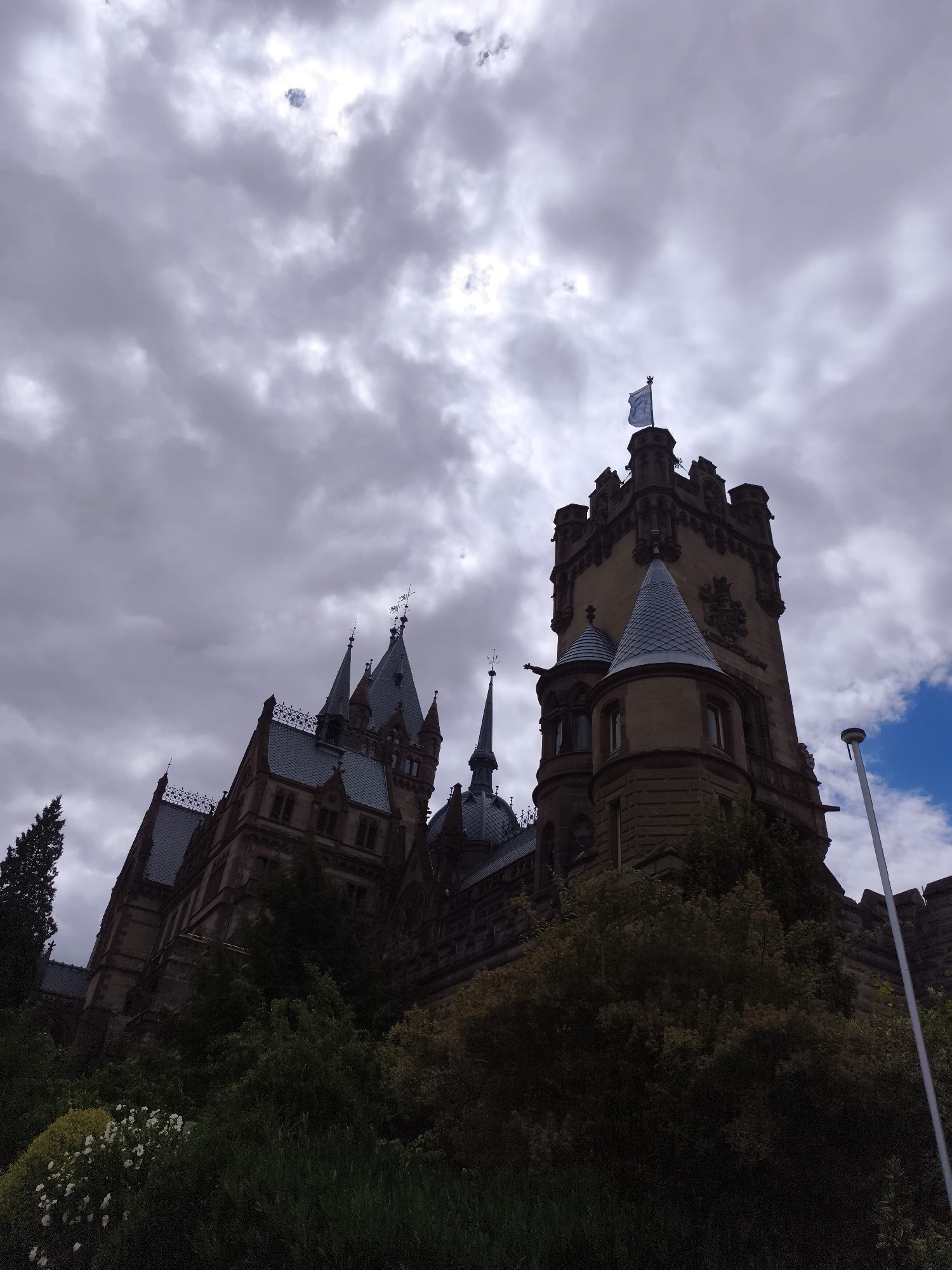 The image shows a dramatic, low-angle view of Drachenburg Castle, a historic landmark situated on the Drachenfels hill in Königswinter, Germany. The castle, constructed of dark stone, features multiple towers with conical roofs and crenellated battlements. A prominent tower on the right flies a light blue flag with a white emblem. The castle is partially obscured by lush green trees and foliage in the foreground, with some white flowers visible on the left. The sky above is filled with heavy, dramatic clouds, with a few patches of brighter sky and a sliver of blue sky visible on the far right. A modern white lamppost stands to the right of the main tower. The overall impression is one of grandeur and a slightly ominous, overcast atmosphere.