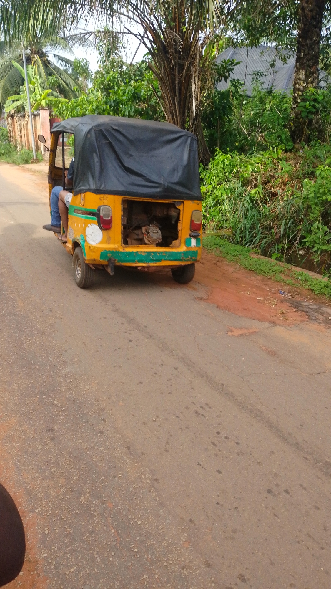 The image depicts a yellow and black "keke", a three-wheeled motorized vehicle, commonly used for public transportation in Nigeria, on a paved road. The keke is missing its rear door and appears to be carrying cargo. The location appears to be in a tropical area, with lush green vegetation on both sides of the road. The pavement has a reddish-brown hue, indicative of the local soil. The photograph was taken in Enugwu-Ukwu, Nigeria. The road is fairly narrow and appears to be in a residential area.