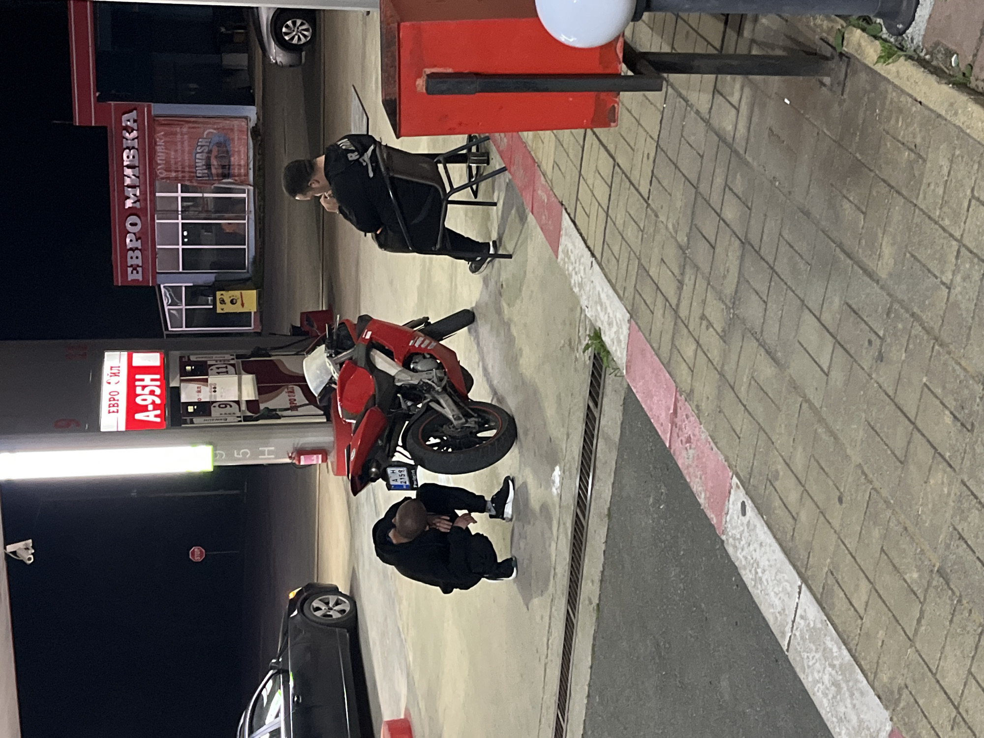 The image shows a scene at a gas station in Yabalchevo, Bulgaria. There are two individuals present. One is sitting on a chair near a red trash can, wearing dark clothing and appearing to be occupied with something in their hands. The other individual is kneeling near a red motorcycle, seeming to be either inspecting or adjusting something on it. The gas station has a sign that says "Евро Мивка" which translates to "Euro Wash", and another with gas prices. A car is parked nearby, partially visible. The setting suggests it is nighttime or late evening.
