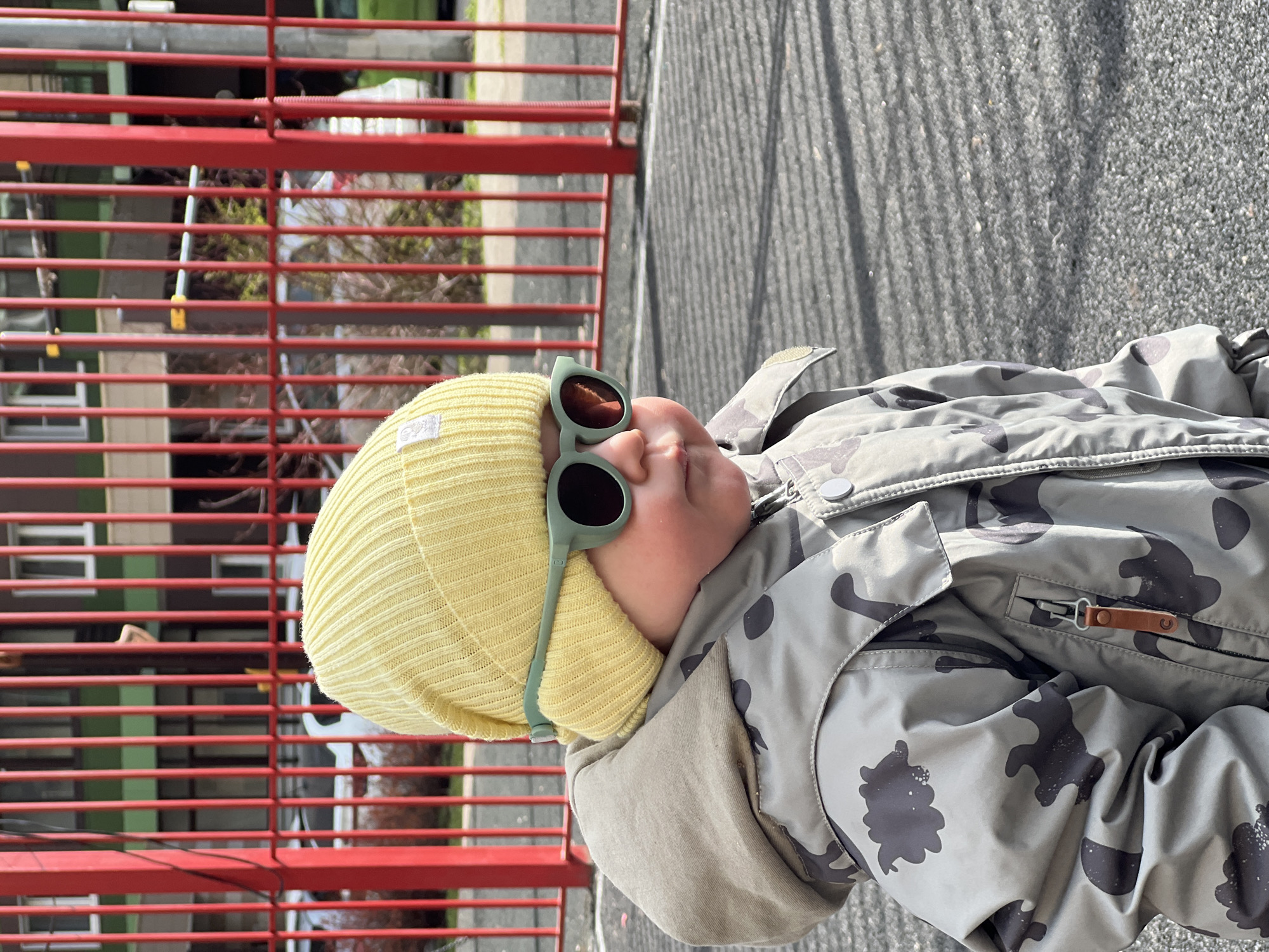 A child, wearing a yellow beanie, round green sunglasses, and a camouflage jacket, is positioned next to a red metal structure. The surface visible in the background is gray with patterns suggesting an outdoor environment. The location is Khimki, Russia.