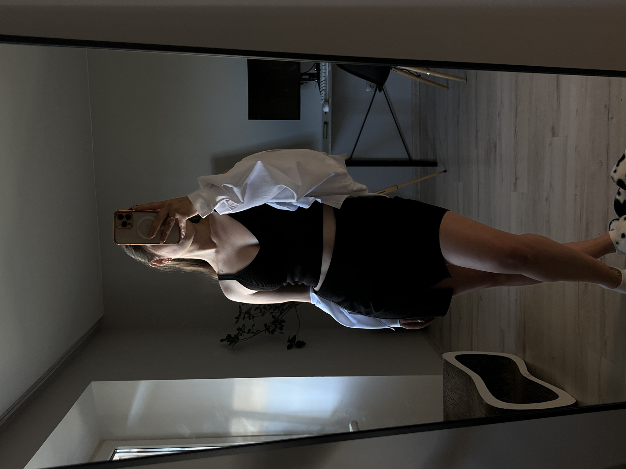 A young woman stands in front of a mirror, taking a selfie. She is wearing a black crop top, a black skirt, and a white open shirt draped over her shoulders. The woman has long brown hair and is holding a phone to take the photo. The reflection in the mirror shows a room with a window on the left, a plant in the background, and a desk with a computer monitor. The floor is wooden. The woman is wearing dalmatian print slippers. This image was taken in Ternopil, Ukraine.