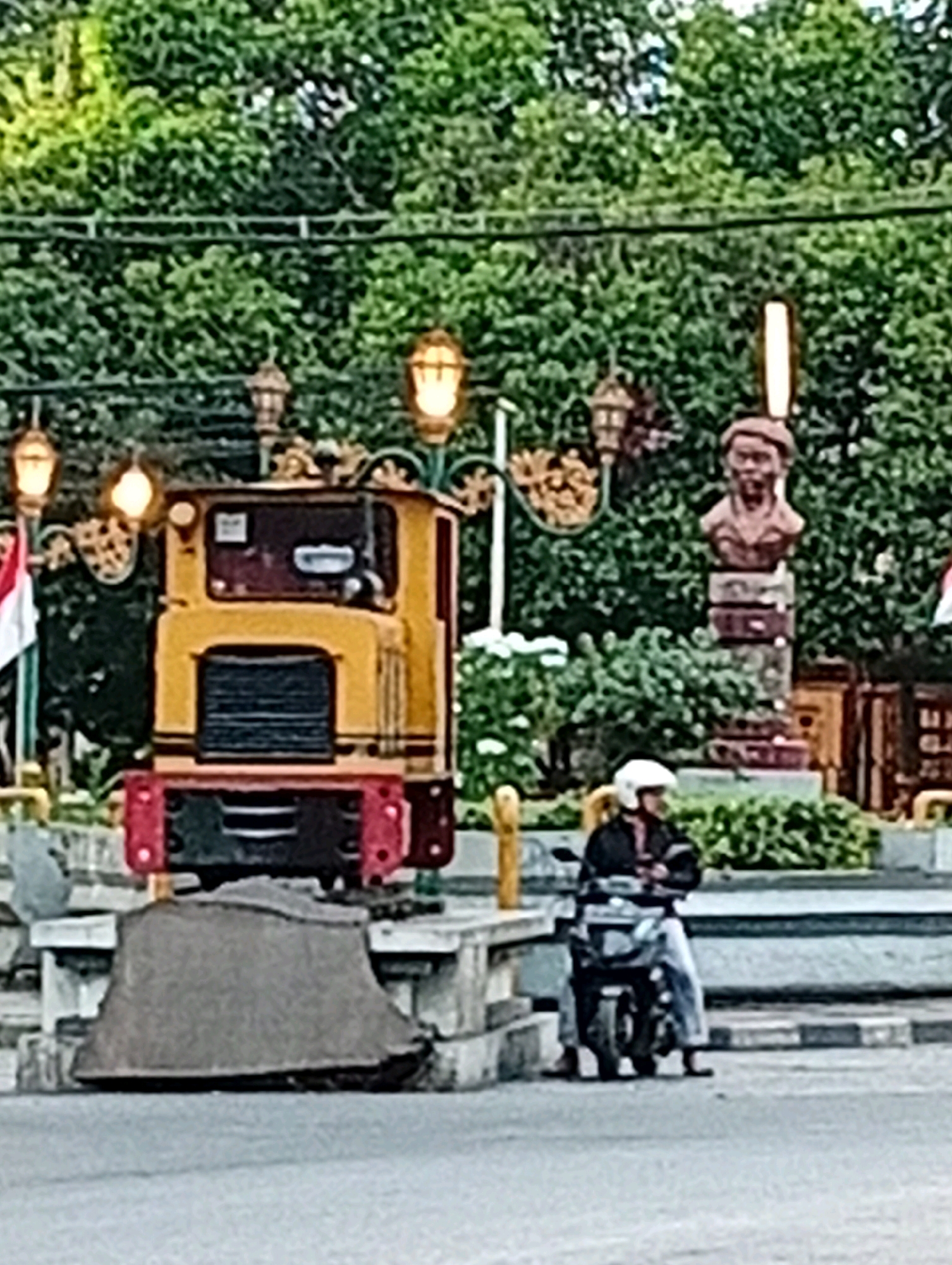 The image captures a scene in Malang, Indonesia, featuring a small, yellow, train-like structure with red accents, likely a decorative element or a miniature ride. To the right of the train, a bust of a man stands on a pedestal, with a lamppost nearby. In the foreground, a person wearing a helmet and a dark jacket is seated on a motorcycle. The environment appears to be an urban outdoor setting with lush green trees in the background. The lighting suggests it is daytime, possibly late afternoon or early evening, with soft light illuminating the scene. The overall impression is that of a public space, potentially a park or a square in the city.