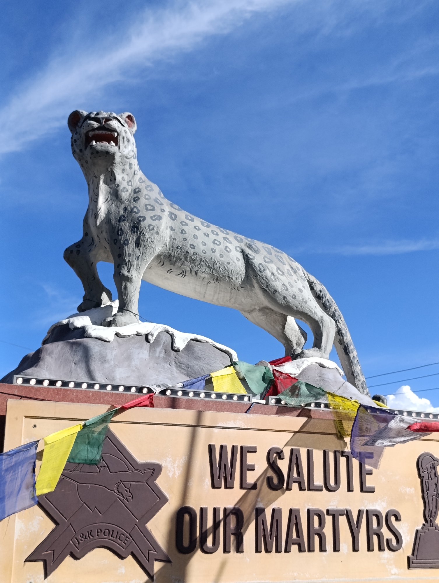 A low-angle medium shot captures a snow leopard statue with its mouth open in a roar, positioned atop a rocky, snow-dusted base. The statue is predominantly gray with black spots, and a black rope is tied around its front paws. Below the statue, a light brown sign displays "WE SALUTE OUR MARTYRS" in large, dark brown embossed lettering. To the left of the text is a crest with "J&K POLICE" written below it, also embossed. Prayer flags in multiple colors (blue, yellow, green, red, white) are strung across the top of the sign and around the base of the statue. The background consists of a bright blue sky with scattered white clouds. The scene is located at the gateway to Leh, India, likely a memorial site given the signage. There are no people present in the image.