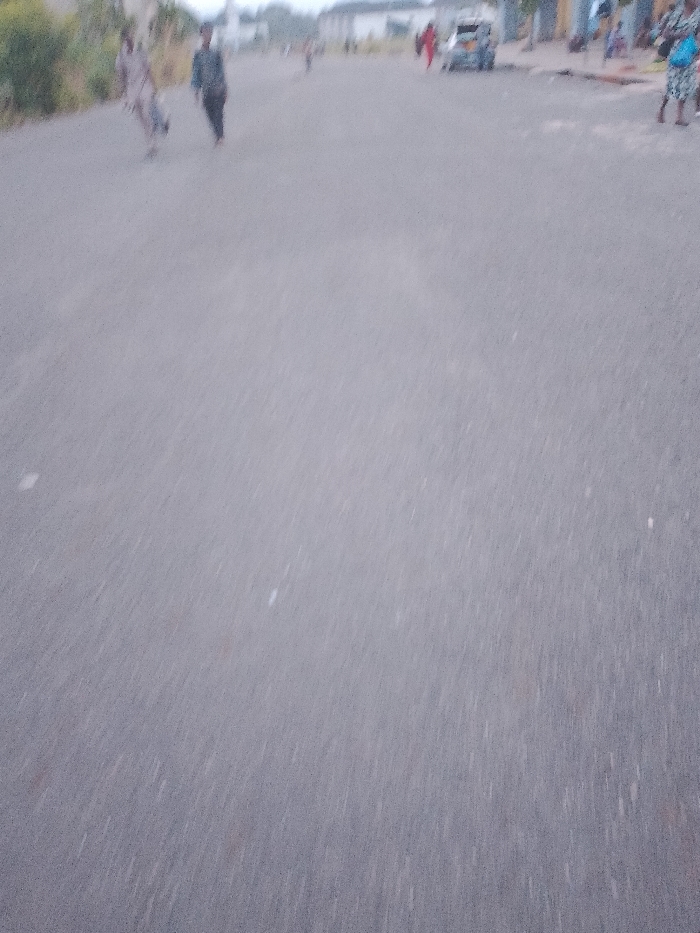 The image depicts a street in Ogboko, Nigeria. The road surface is asphalt. People are present on the street, including individuals walking and standing. A vehicle is parked in the distance. Buildings line the street. The scene appears to be taken during daylight hours.