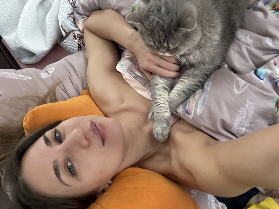 A close-up, high-angle selfie photograph depicts a young adult female lying in a bed next to a grey domestic cat. The female, possessing brown hair and light-colored eyes, is positioned on her left side with her head resting on an orange cylindrical pillow. She is partially covered by a light purple duvet adorned with a pattern of stylized colorful feathers. Her bare right shoulder and arm are visible, and her right hand gently rests upon the cat's back. The cat, appearing to be a grey-furred breed such as a British Shorthair, lies on its left side, partially beneath the duvet, with its head turned away from the camera and towards the female's chest. Additional bedding, including a light blue textured blanket, is visible in the background. The scene is set within a domestic bedroom environment. No specific visual information within the image identifies its geographical location in Saint Petersburg, Russia, or any other specific city.