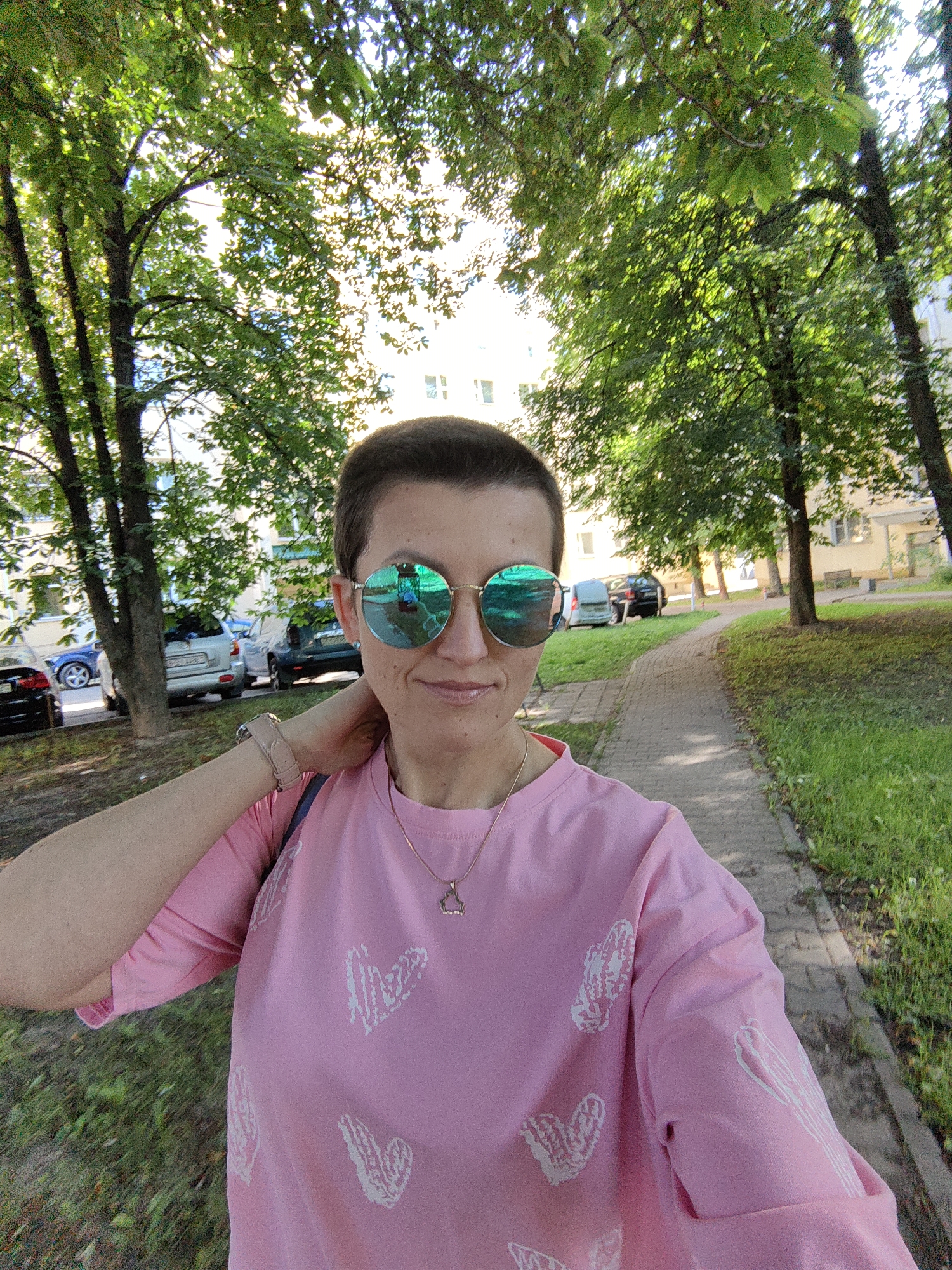 A woman with short brown hair and green mirrored sunglasses is taking a selfie outdoors on a paved path lined with grass and trees. She is wearing a pink t-shirt with white heart designs and a thin gold necklace. Her right arm is bent behind her head. In the background, parked cars and apartment buildings are visible. The scene appears to be in a residential area of Minsk, Belarus, based on the architecture and vegetation.