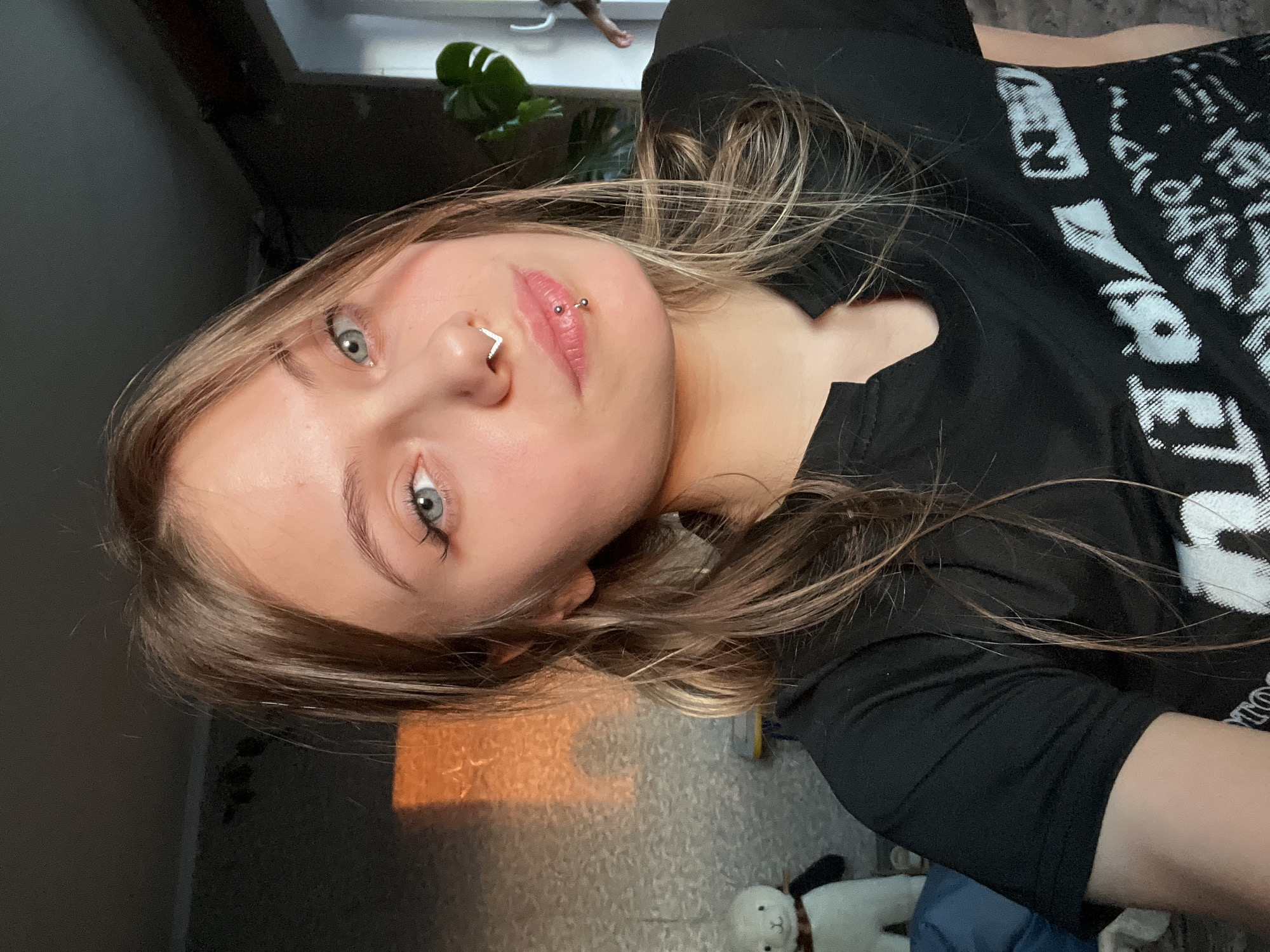 This media file presents a close-up, angled selfie of a young individual indoors. The subject has light skin, striking light blue-grey eyes, and long, layered light brown hair with blonde highlights. They feature a V-shaped septum piercing and a barbell-style philtrum piercing (medusa). Their gaze is direct, with a neutral expression.
The person is wearing a black short-sleeved t-shirt with a prominent white, distressed graphic design or text across the chest, though no specific words are clearly readable. The setting appears to be a room within a home, well-lit by natural daylight streaming from a window in the background. A large-leaf green plant is partially visible near the window. Sunlight creates patterns and reflections on a light-colored, patterned floor that extends across the bottom right of the frame. A small, white plush toy, possibly a Miffy doll, is visible on the floor in the bottom right corner.
The image is taken from an elevated or tilted perspective, suggesting the person may be reclining. The natural light indicates it is daytime. The overall atmosphere is casual and personal, typical of a domestic environment. No specific visual cues confirm the geographical location of Фоки, Russia.