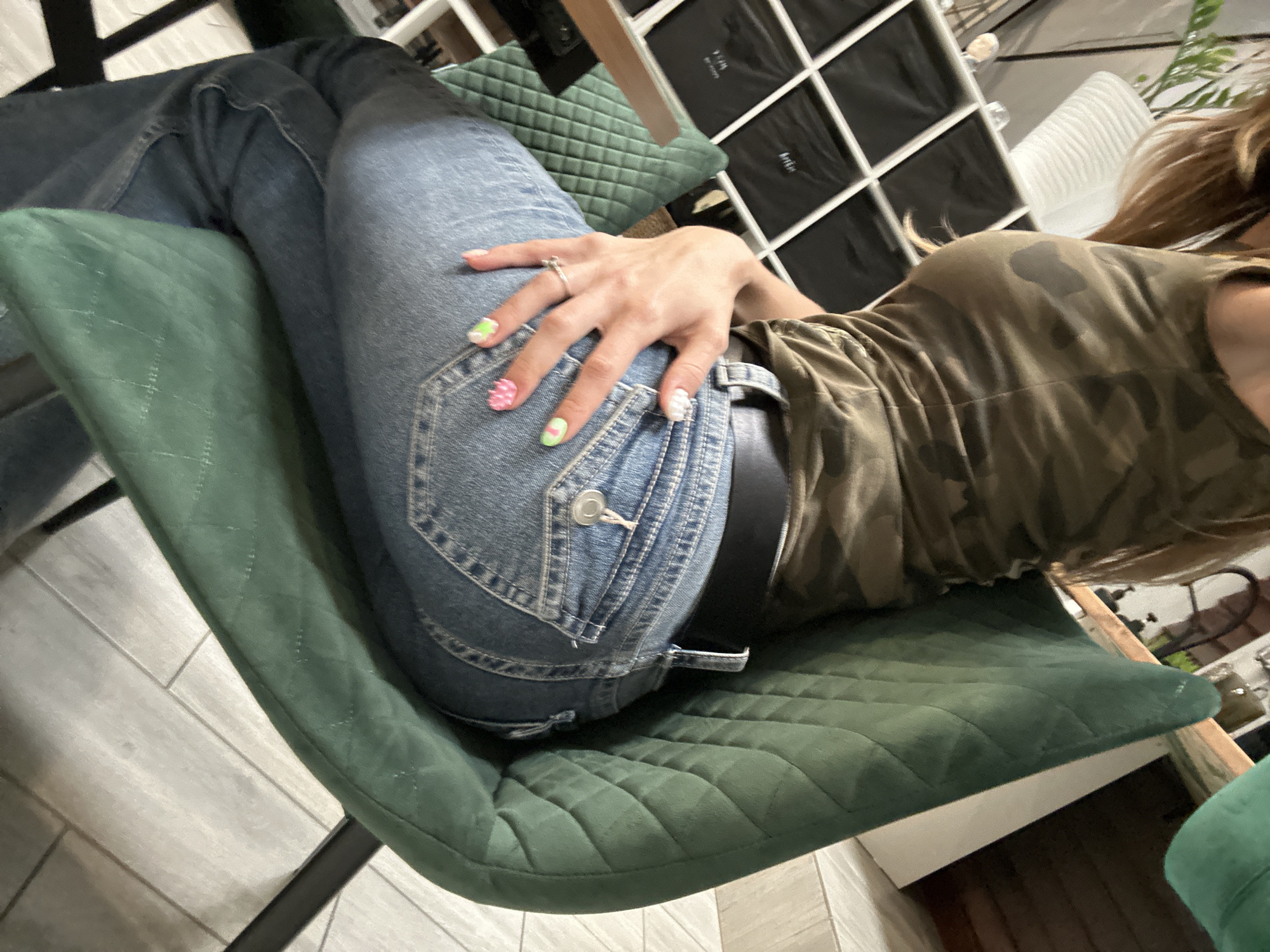 A person is sitting in a green quilted chair, wearing a camouflage-patterned tank top and blue jeans. Their right hand is resting on their left hip, with fingernails painted in various colorful designs. The background shows a white shelving unit filled with black bins, and a potted plant. The lighting suggests an indoor setting. The image does not contain any elements that can be used to determine its location in Moscow, Russia.