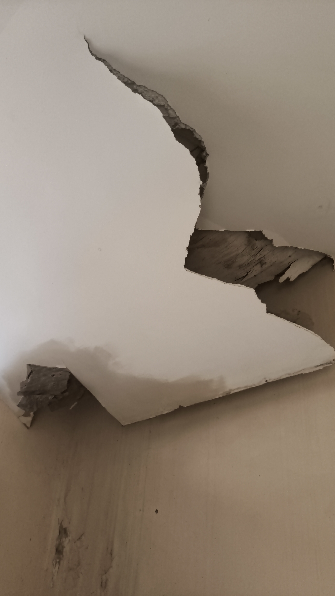 The image depicts significant damage to a white ceiling, likely caused by water intrusion. A large section of the ceiling plaster has cracked and peeled away, exposing wooden beams or structural elements beneath. The exposed material appears aged and possibly damp. The wall below the damaged ceiling is a neutral, light brown color and shows water stains and discoloration, suggesting a history of leaks. The scene appears to be an interior residential or commercial space. The location is a property in South Jakarta, Indonesia.
