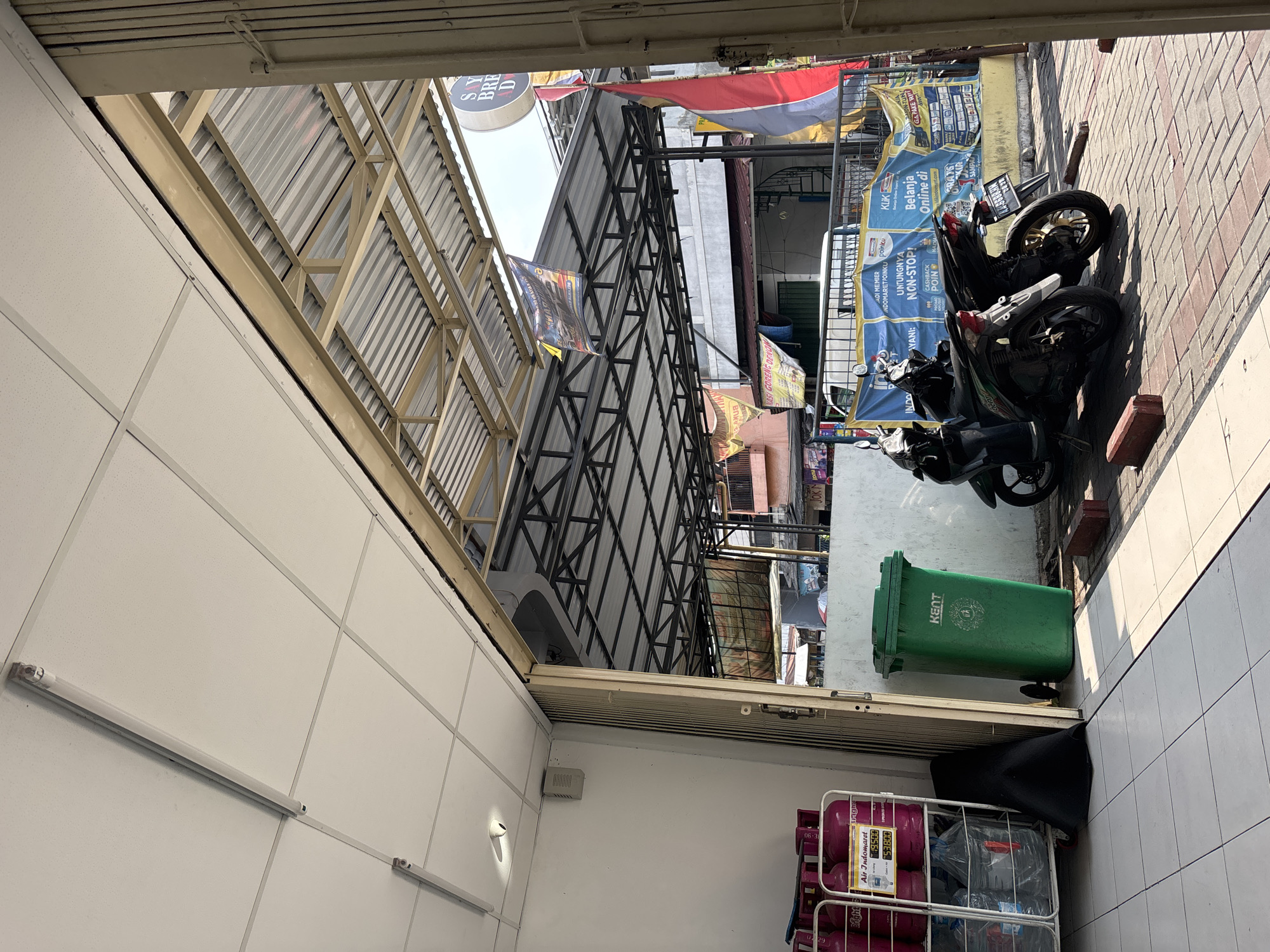 The image depicts the exterior of a small retail establishment in North Jakarta, Indonesia, likely a convenience store or minimarket. The storefront is open, with a large metal accordion door partially retracted. Inside, a white tiled floor leads to shelves stocked with pink gas cylinders, some with water bottles on a rack. To the right of the entrance, a large green plastic trash bin labeled "KENT" stands prominently.
Outside the establishment, the ground is paved with interlocking red and gray bricks. Two parked scooters, a dark green and a black model, are positioned in front of a blue and yellow banner advertising various services, including "Indomaret," "Poinku," and "KLIK." The banner also features multiple logos of brands and payment methods. Red brick debris is scattered on the ground near the scooters. A corrugated metal roof structure extends from the building, supported by a metal frame, and several flags and banners are visible hanging from it, including a red, white, and blue flag and a circular sign with text that appears to read "SAY BRE D." The overall scene suggests a busy commercial street environment.