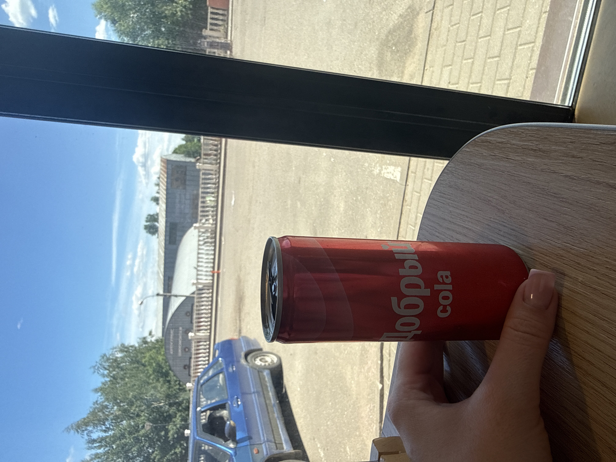 A hand holds a red aluminum can of Dobriy cola on a wooden table. The can is partially visible from the side and is positioned in the foreground. In the background, a blue car is parked on a street with a white railing and some buildings. The scene appears to be outdoors on a sunny day, with a clear blue sky and trees visible. The location is identified as Golitsyno, Russia.