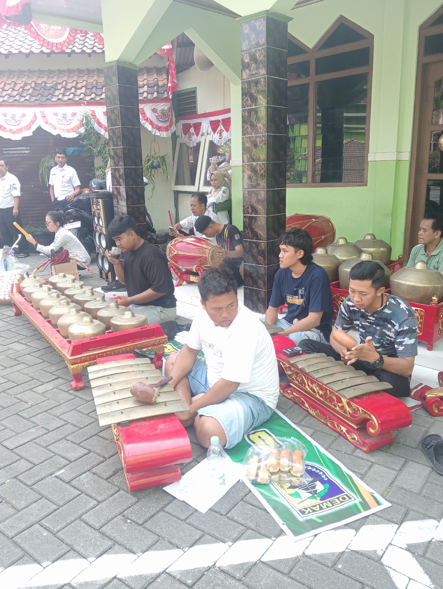 The image captures an outdoor scene in Demak, Indonesia, likely in front of a building with a traditional Indonesian architectural style. Several people are gathered, some playing musical instruments that appear to be part of a gamelan ensemble. Two main instruments, a metallophone with bronze keys and a large drum, are prominently displayed.
In the foreground, a young man in a white t-shirt and blue shorts is seated, looking down. To his right, another young man in a camouflage t-shirt is holding a mallet, seemingly ready to play. In the middle ground, a man in a black t-shirt is drinking from a small cup while seated near a metallophone. A woman in a white shirt and glasses is holding a yellow stick, positioned as if to play an instrument or give instructions. Further back, several other individuals are visible, some watching the musicians, others standing.
The environment suggests a public space, possibly a courtyard or a plaza, with patterned brick paving. Red and white decorative banners are hanging from the building, hinting at a festive occasion or a national celebration. The building itself has green walls and wooden-framed windows. A few plastic bottles and bags of snacks are scattered on the ground near the musical instruments. A banner with the word "DEMAK" and a coat of arms is spread on the ground. The lighting suggests it is daytime, with some shadows indicating direct sunlight.
