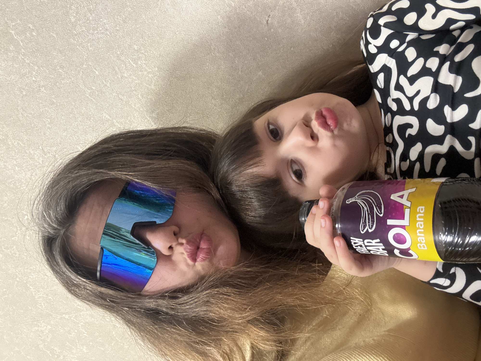 The image captures two Caucasian females, an adult and a child, posing in an indoor setting in Chaikovsky, Russia. The adult female, positioned to the upper left, displays long light-brown hair with visible gray strands and wears a mustard-yellow long-sleeved garment. She is equipped with large, wrap-around style sunglasses featuring iridescent blue and purple reflective lenses. Her lips are conspicuously pursed. The younger female child, positioned to the lower right, has dark brown hair with bangs and wears a black long-sleeved top with a white abstract pattern. Her lips are also pursed, mirroring the adult's expression. The child holds a clear plastic bottle with a black screw-cap in her right hand. The bottle's label is purple and yellow, clearly indicating "FRESH BAR COLA Banana" alongside a graphic of two bananas, and contains a dark brown liquid. Both subjects are looking directly towards the camera. The background consists of a light-colored, textured wall.