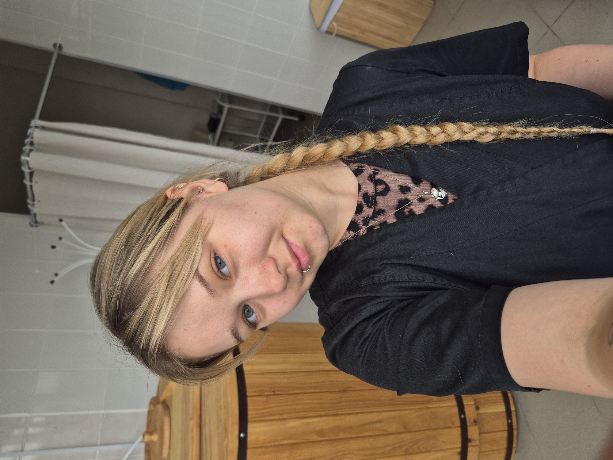 A young Caucasian woman with blonde hair braided to her right side and blue eyes is looking at the camera. She is wearing a black scrub top over a leopard-print shirt, and a silver necklace with a pendant. She has piercings on her left ear and a ring in her lower lip. She is standing in front of a wooden barrel. In the background, a shower curtain and tiled walls are visible. The scene appears to be in a spa or sauna facility located in Chaikovsky, Russia.