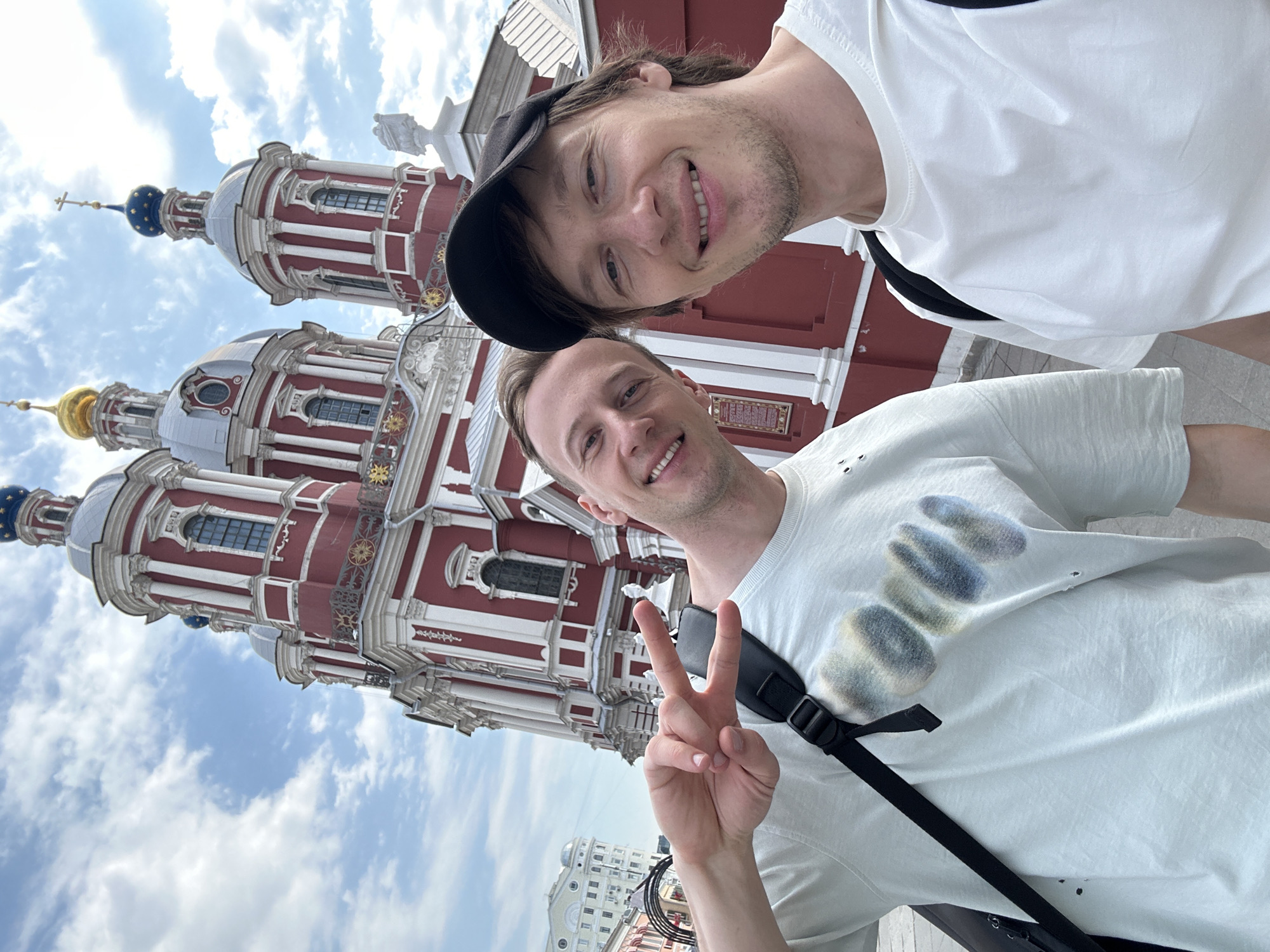 Two men are posing for a selfie. One wears a white t-shirt with a black strap across his chest and is making a peace sign with his right hand. The other man is wearing a white t-shirt and a black cap. Behind the men is the Donskoy Monastery in Moscow, Russia. The building is red and white with multiple domes and spires. The sky is blue with some clouds.