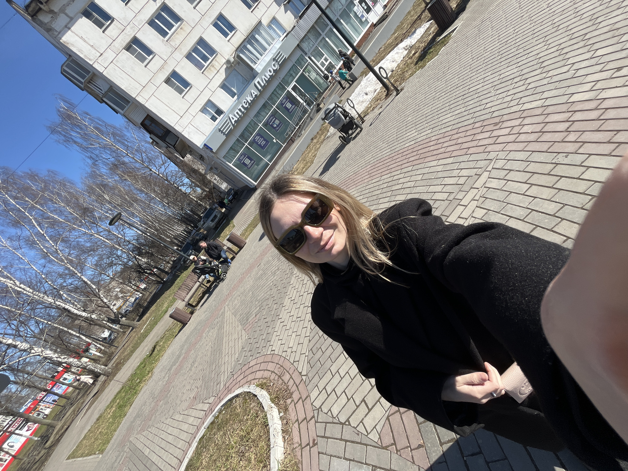 A woman wearing a black coat and sunglasses is taking a selfie on a sunny day in Chaikovsky, Russia. The background includes a building labeled "Аптека Плюс" (Pharmacy Plus), a sidewalk with a circular brick design, bare trees, parked cars, benches, and a street. There are people with a stroller in the background. The woman is holding her hands together near the bottom of the frame.