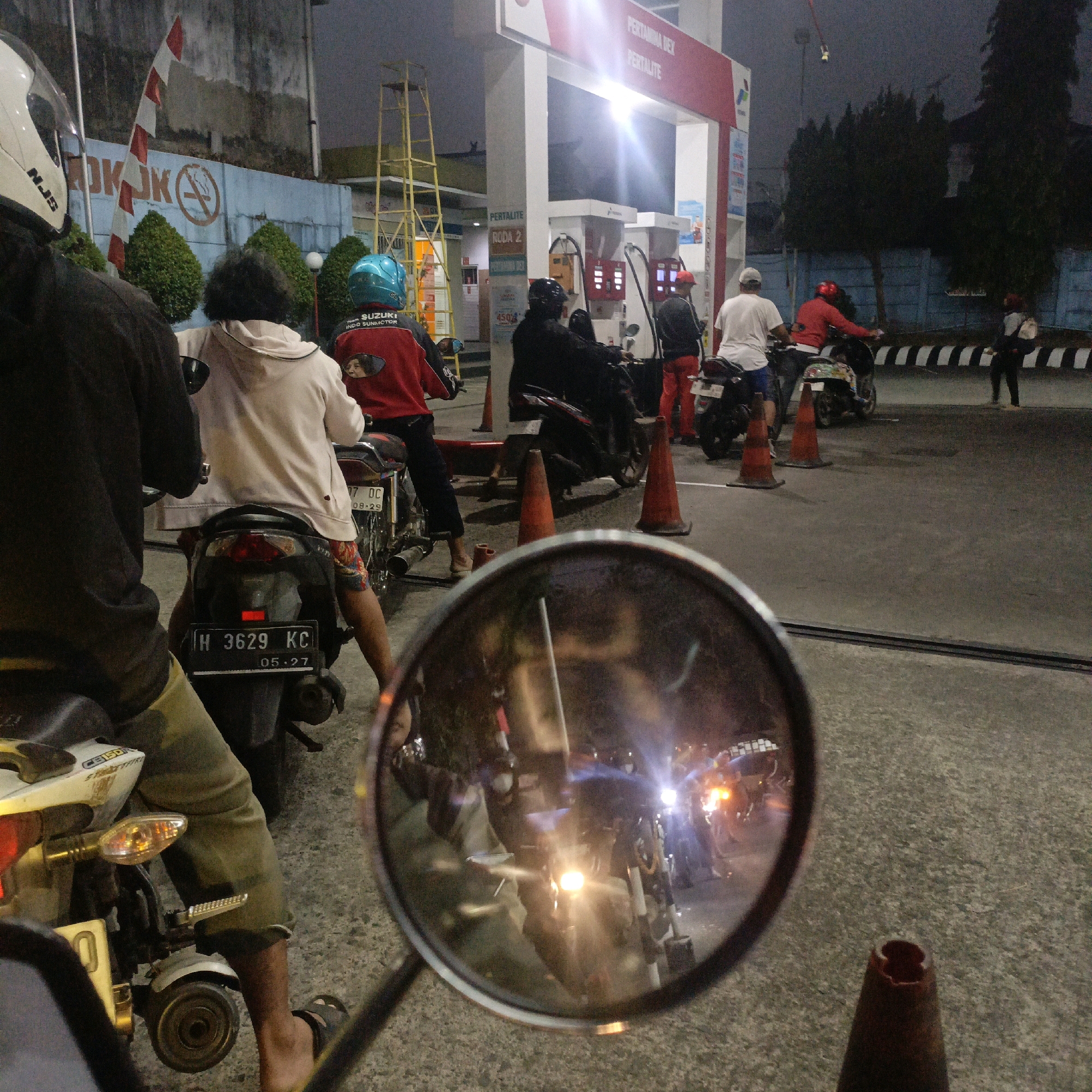 The image depicts a nighttime scene at a Pertamina gas station in Salatiga, Indonesia, likely along a busy road. In the foreground, a person wearing a white helmet with "NJB" visible is seated on a motorcycle. To their right, another person in a light-colored hoodie and patterned shorts is also on a motorcycle. Behind them, a person in a red jacket with a blue helmet is also on a motorcycle. In the background, the Pertamina station's canopy with red and white signage is illuminated. Several motorcycles are lined up at the gas pumps, with attendants in red and white uniforms visible. Other individuals are also present, either on motorcycles or standing. Orange traffic cones are placed around the area. The ground is paved asphalt. The atmosphere suggests a typical urban evening at a gas station, with a sense of ongoing activity.