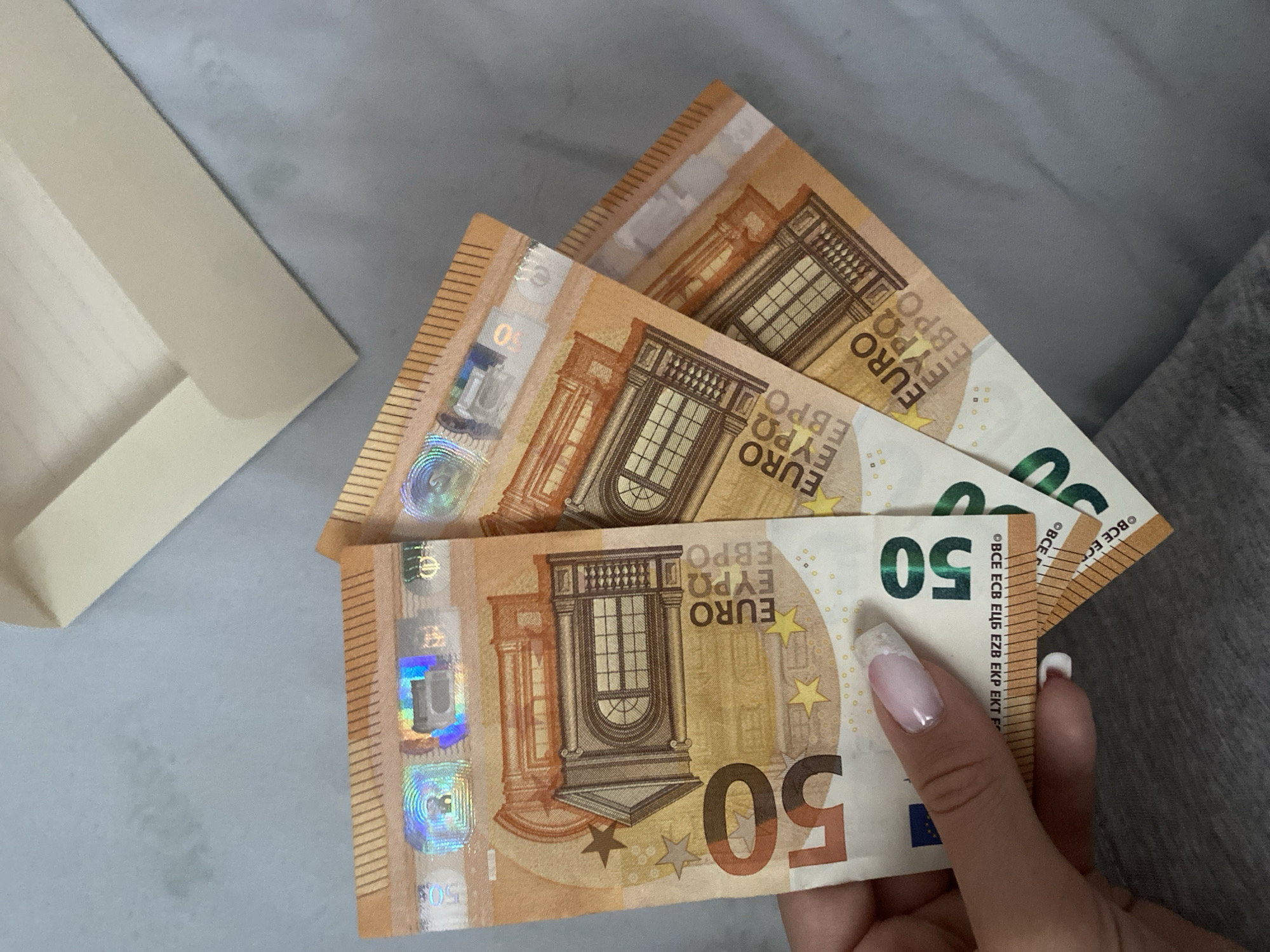 An overhead shot captures a hand holding three fanned-out 50 Euro banknotes against a light grey background. The banknotes, predominantly orange and brown in color, display architectural designs of windows and columns, along with holographic security features. The hand, with manicured nails featuring a white French tip design, is holding the currency in the lower right portion of the frame.
To the left of the banknotes, a corner of a light beige or cream-colored envelope or folded paper is visible on the grey surface. The grey surface itself appears slightly textured, possibly a table or a flat fabric. Further to the right, a darker grey, soft-looking material, perhaps part of clothing or bedding, can be seen.
The activity depicted is simply a person holding Euro currency. No specific event or strong emotions are discernible. The setting appears to be an indoor environment in Blagoevgrad, Bulgaria.
Visible text on the banknotes includes "50", "EURO", "ΕΥΡΩ", "ЕВРО", and security text like "BCE ECB EZB EKP EKT F".
