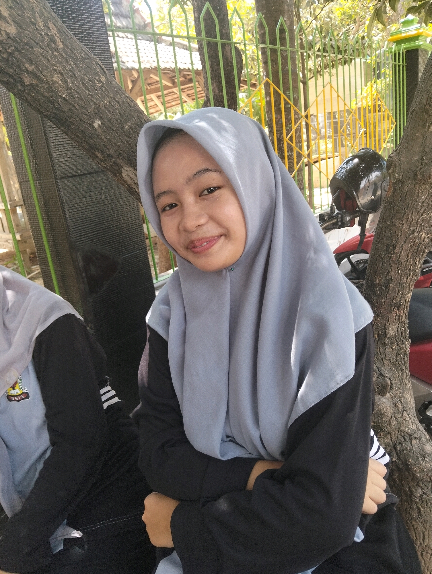 In a sunlit outdoor setting in Paciran, Indonesia, a young woman wearing a light blue hijab and a black long-sleeved shirt is captured in a close-up portrait. She is seated, with her arms crossed, and her expression is a gentle smile. Behind her, a tree trunk dominates the left side of the frame, while a fence with yellow and green accents and a red motorcycle with a black helmet are visible in the background. Another individual, also wearing a hijab and a blue and black outfit, is partially visible on the left. The environment suggests a casual, possibly school-related, outdoor area with natural foliage.