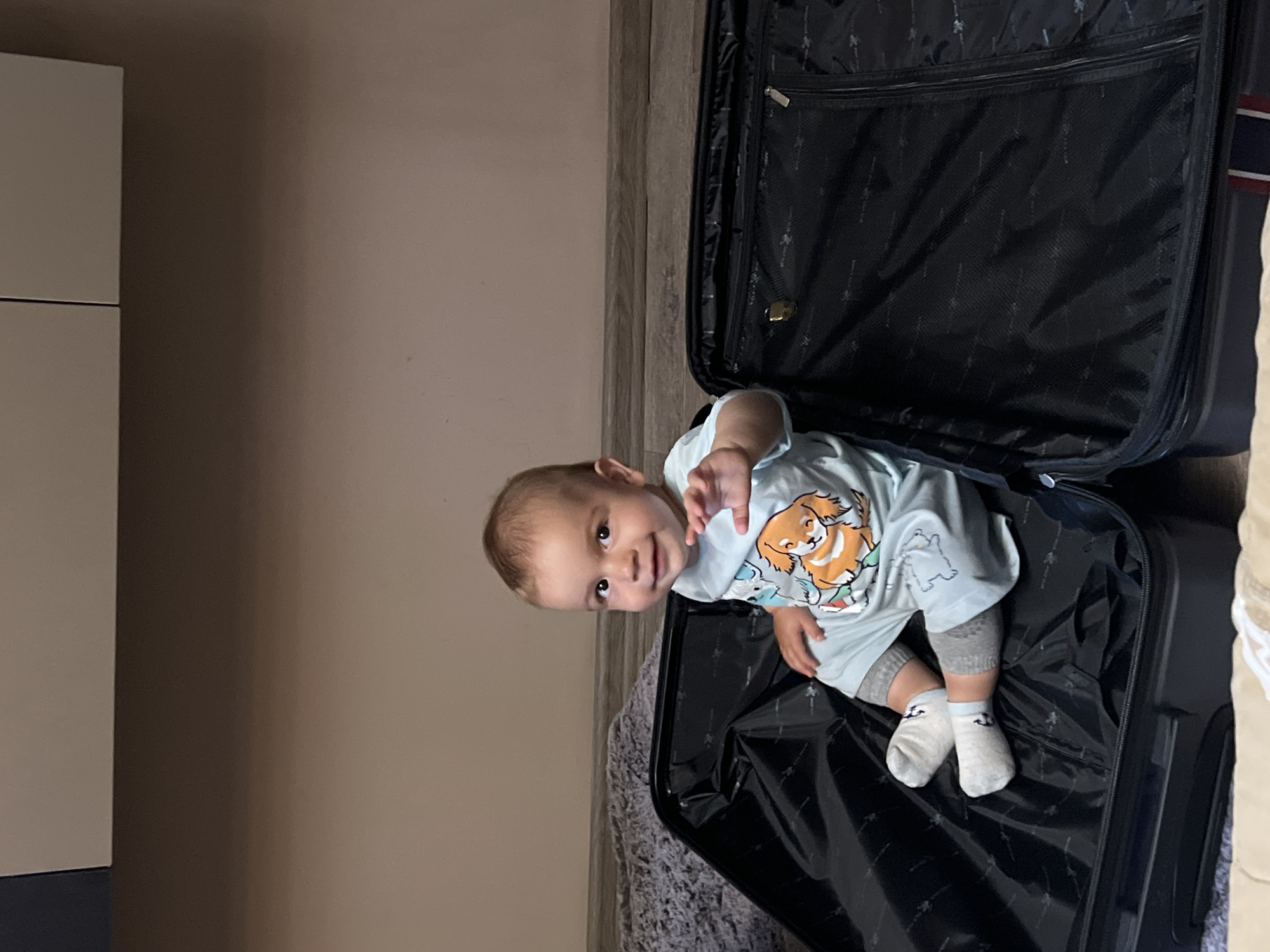 A young child is playfully seated inside an open, black hard-shell suitcase in an indoor setting in Burgas, Bulgaria. The child, with light brown hair and dark eyes, is smiling broadly and looking directly at the camera, with one hand slightly raised as if waving. They are wearing a light blue t-shirt with a cartoon graphic of a brown and white dog, grey shorts or leggings, and white socks with a small black anchor motif.
The large suitcase lies open on what appears to be a grey, shaggy rug or carpet. Its black interior lining features a subtle, repeating pattern, and the propped-open lid reveals a zippered mesh compartment. To the left of the image, a segment of a white cabinet or dresser is visible against a light beige wall. A wooden panel can also be seen vertically behind the suitcase on the right side of the frame. The lighting suggests an indoor environment. The overall mood is cheerful, capturing a moment of playful exploration.