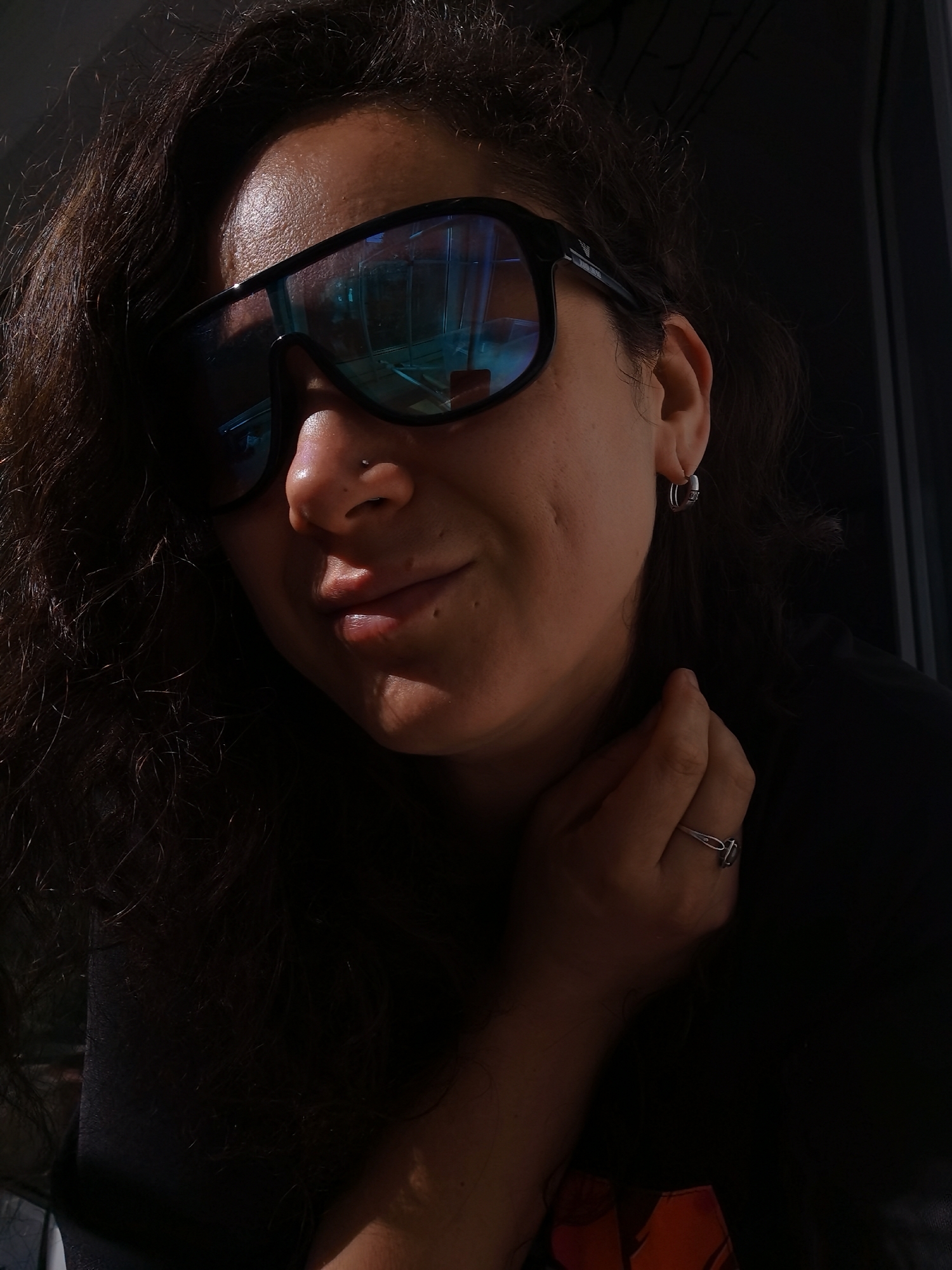 A close-up, waist-up shot shows a person wearing large, black-framed sunglasses with blue lenses. The person has dark, curly hair and is wearing a simple silver hoop earring in their left ear and a silver ring on their right ring finger. The person's right hand is resting on their neck. The lighting suggests the sun is to the left of the frame, casting a warm glow on the person's face and illuminating one side of their hair. The background is dark and out of focus, with the reflection of windows visible in the sunglasses. There is no indication of the specific location within Chaikovsky, Russia, or any outdoor elements that would suggest a city environment.