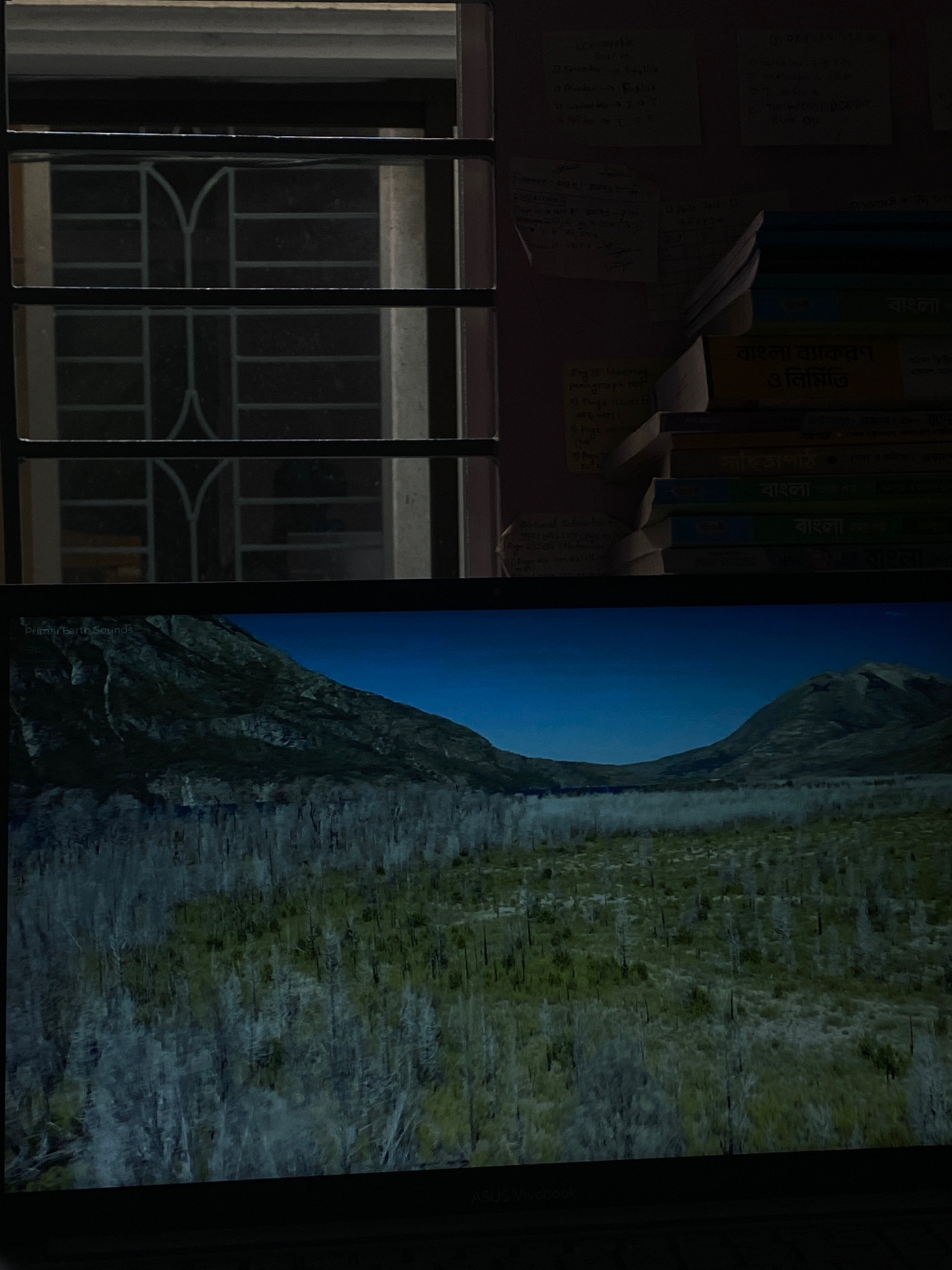 The image is a split view, with the top portion showing a window with a metal grate and light filtering through, illuminating a wall with stacked books and notes. The books appear to have Bengali text on their spines. The bottom portion displays a landscape scene on a screen, featuring rolling hills, a body of water, and sparse vegetation. The text overlay on the bottom left of the screen reads "Primal Earth Sounds." The bottom right of the screen shows faint text that appears to be "ASUS Vivobook". The image depicts a domestic indoor setting combined with a digital display of a natural landscape, with no people visible. The location is not specified in the image itself, but the request mentions Hemayetpur, Bangladesh, which is not visually evident in the provided image content.