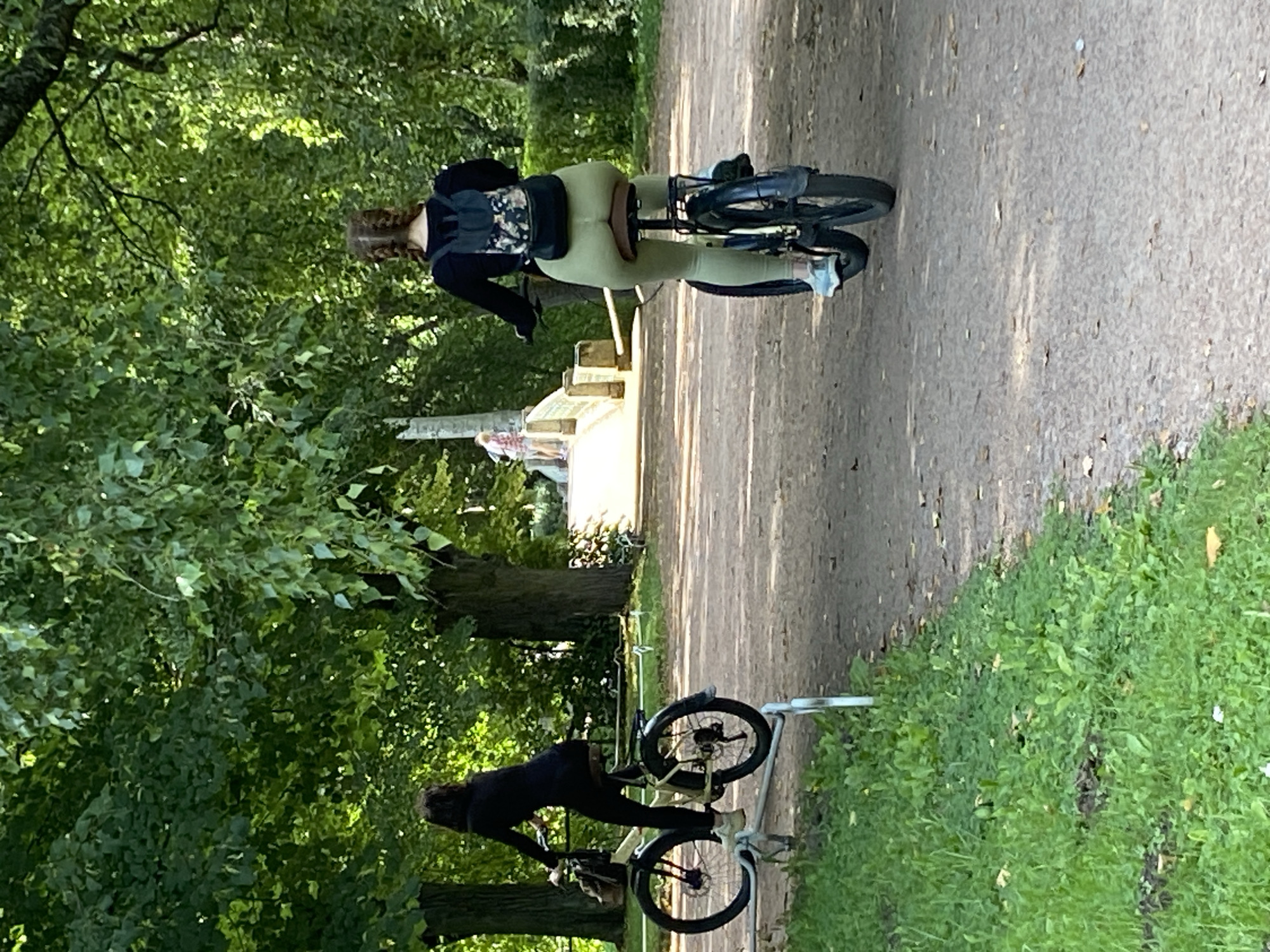 The image depicts a park scene in Pavlovsk, Russia, with two individuals on bicycles on a gravel path. In the foreground and slightly to the left, a woman with dark hair is riding a light-colored bicycle. She is wearing dark clothing. The bicycle has a basket on the front. To the right, another woman with braided hair is riding a dark-colored bicycle. She is wearing a black long-sleeved shirt and light green leggings. She has a patterned backpack on her back. In the background, a bridge with ornate railings is visible, with a few people walking on it. The environment is lush with green trees and foliage, suggesting a park setting. Sunlight filters through the trees, casting shadows on the path.