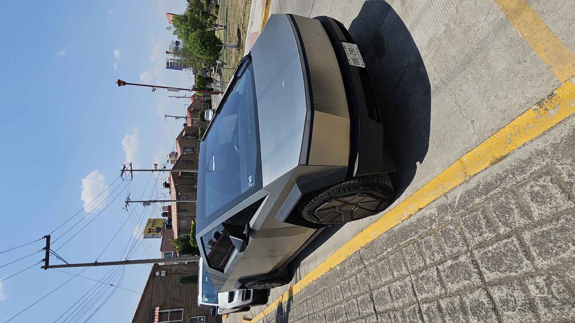 A gray Tesla Cybertruck is parked on a concrete street with yellow painted parking lines in San Bernardino Tlaxcalancingo, Mexico. The car is angled in the parking space with a license plate visible on the back. Adjacent is a white vehicle with visible wheel and tire. The street is paved with rectangular brick stones. Behind the Cybertruck, there are residential buildings and power lines. The sky is blue with sparse clouds. A red street light and green trees are visible in the background.