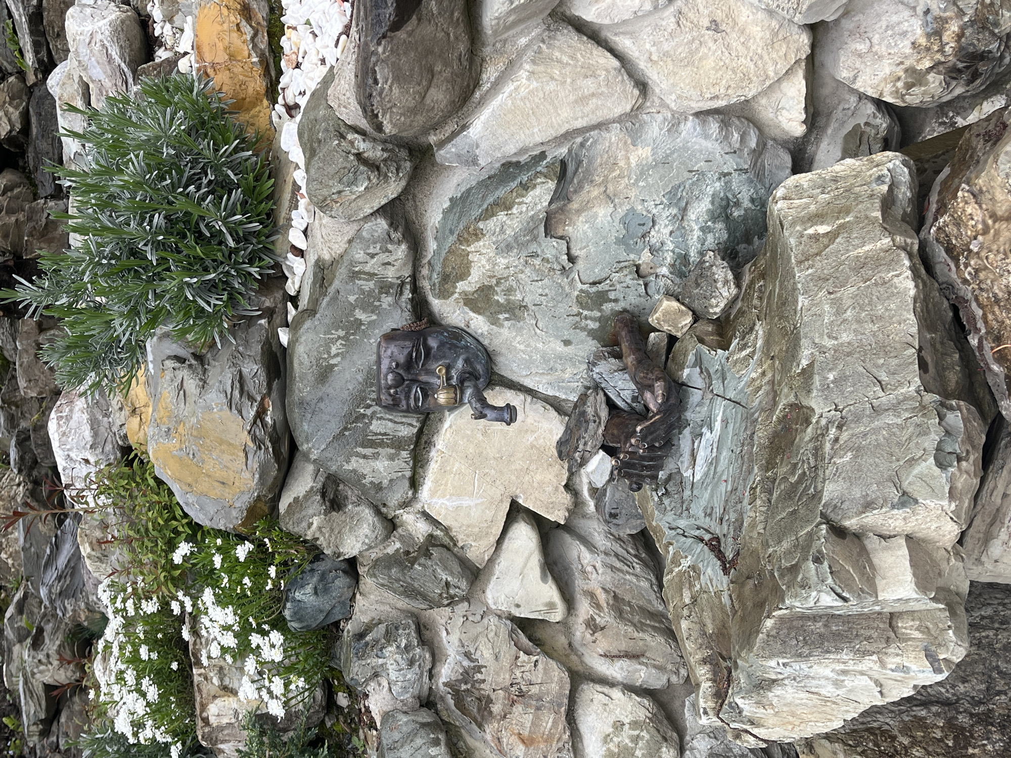 The image shows a rock garden, constructed from various gray and tan stones of different sizes and shapes. Patches of green plants, including rosemary and white flowering ground cover, are interspersed between the stones. Mounted on the rock wall are two dark-colored metal sculptures: one appears to be a human face with mechanical components, and the other appears to be hands grasping something. The scene is situated outdoors in an urban location.