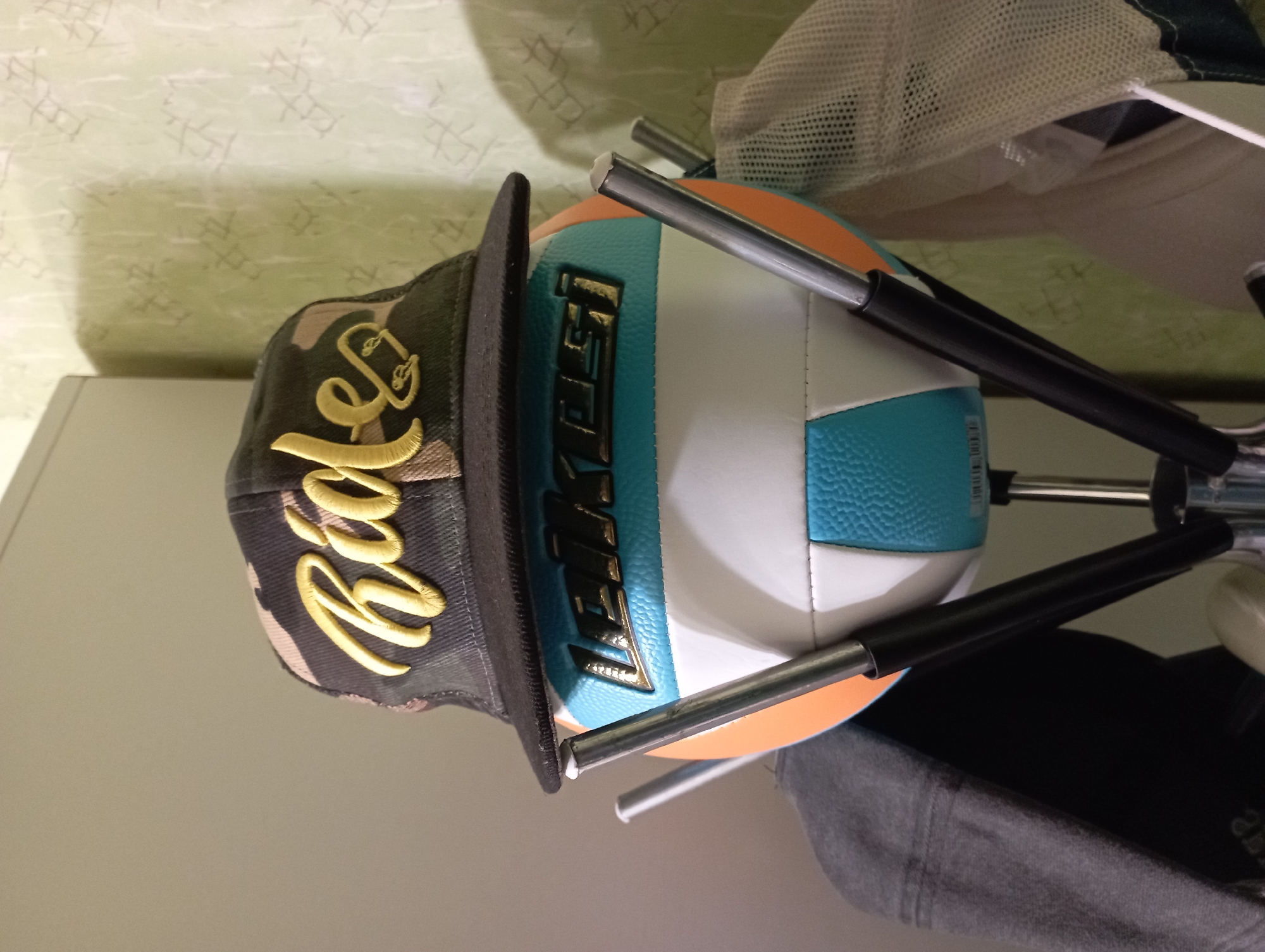 An overhead shot shows a volleyball wearing a camouflage baseball cap with yellow lettering that reads "Rides." The volleyball is orange, white, and blue, with black lettering that reads "LEIKESTU" on the front. The volleyball is mounted on a metal rack with several other items of clothing, including a gray hooded jacket and a white mesh hat. The background is a light green textured wallpaper. The image appears to have been taken indoors, possibly in a storage area or a locker room. There is no indication of the specific location in Blagoveshchensk, Russia.