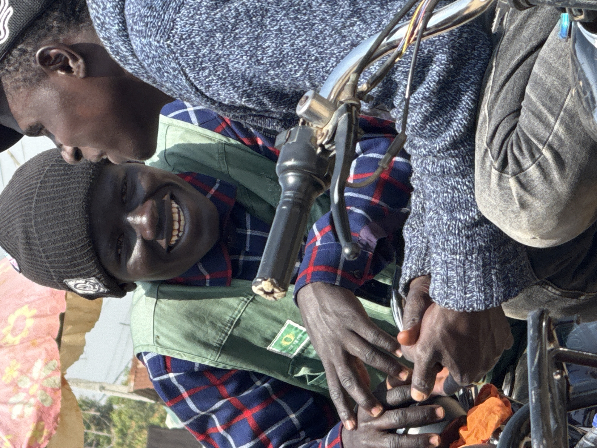 In Abuja, Nigeria, two young men are seated on a motorcycle. The man in the foreground is wearing a dark beanie, a blue, red, and white plaid shirt, and a green vest. He is looking at the camera and smiling, with his hands resting on the motorcycle's handlebars. The man behind him is wearing a dark knitted sweater and looking away from the camera. The motorcycle has chrome handlebars and appears to be in an outdoor setting, possibly a market, as indicated by the patterned canopy in the background. The lighting suggests it is daytime.