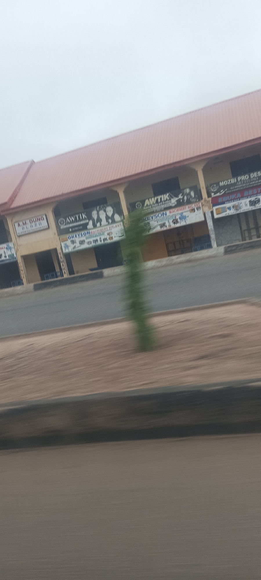 The image captures a commercial building from a moving perspective, indicated by significant motion blur in the foreground and a blurry green plant in the median. The scene appears to be an overcast day.
The primary subject is a long, single-story commercial building with a reddish-brown corrugated metal roof and a light-colored facade, featuring multiple shopfronts. The building's design includes arched or recessed sections over each entrance, supported by columns. In the foreground, there's a paved road, followed by a dirt or sandy median strip, and then the base of the building.
Visible text on various signs identifies the businesses and the plaza:
* On the far left, a prominent sign reads "A.M. DUNG PLAZA" with "RC - 3316..." below it.
* Further right, a black banner features the "AWTIK" logo (a stylized letter A with a crown or similar design) and text that appears to be "Mobile Phone and Laptop Repair & Sales". Below this banner, a sign reads "OKEYSON MACHINERY RIG. ENT." with illustrations of machinery.
* Another section of the building has a similar "AWTIK" banner and below it, "OKEYSON MACHINERY SIG. ENT."
* On the far right, a sign displays "MOZBI PRO DESIGNS" with "ART & STUDIO" underneath. Below this, another sign is partially visible, starting with "KIMUKA BEST...".
The environment suggests an urban or semi-urban area in Jos, Nigeria, given the provided context. No specific human activity is clearly visible, and the shops appear closed or empty. The time of day seems to be a cloudy day, rather than bright sunshine.