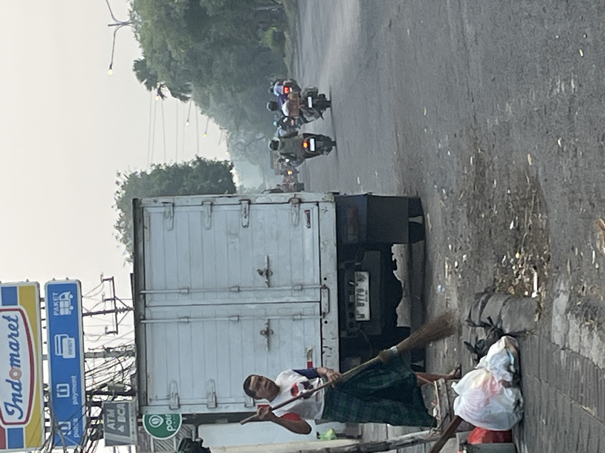 In Kludan, Indonesia, a man with short dark hair, wearing a white t-shirt with red and blue accents and a green checkered sarong, sweeps a dusty road in front of a large white truck. The truck has a white box body and appears to be parked on the side of a busy street. Behind the truck, several motorcycles with riders are moving down the road. The scene is characterized by a hazy sky, suggesting morning or evening light, and a roadside environment with trees and overhead wires. Signs for "Indomaret," "ATM BCA," and various payment services are visible in the background, indicating a commercial area. Trash bags and debris are present on the sidewalk next to the man.