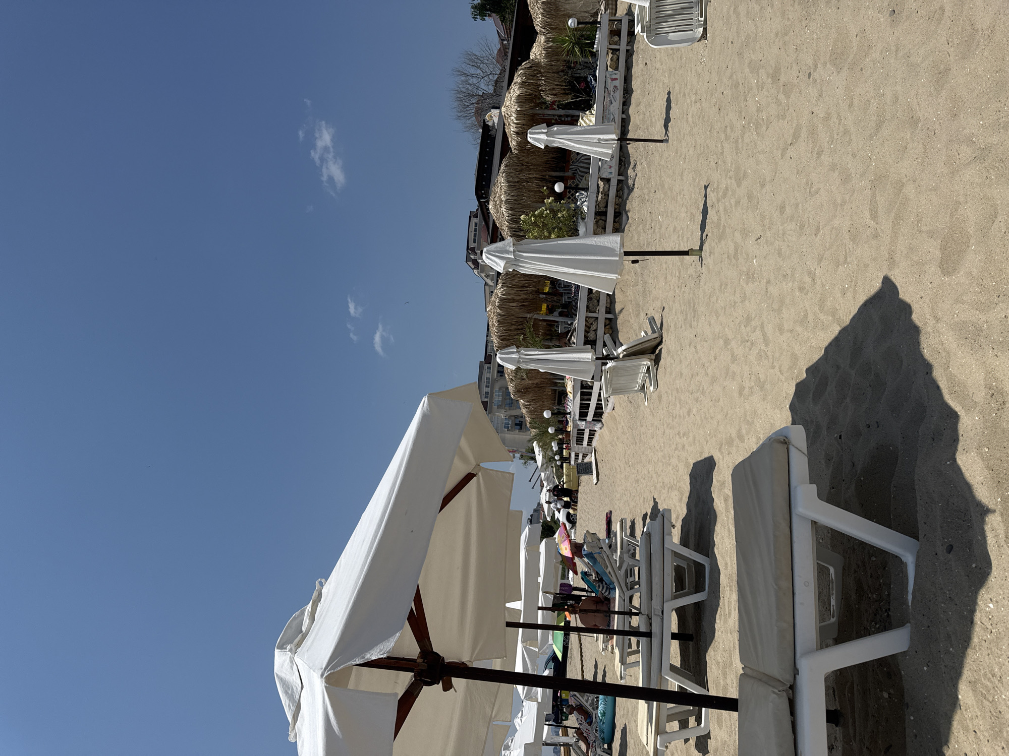 This is a daytime, eye-level, outdoor, medium shot photograph taken on a sandy beach in Sveti Vlas, Bulgaria. The scene depicts a beach resort area with numerous white sun loungers and umbrellas arranged on the sand. In the foreground, several empty white sun loungers are visible, some partially covered by the shadows of large white beach umbrellas.
In the mid-ground, more sun loungers and umbrellas are present, with a few people seated on some of them. A woman is visible sitting on a sun lounger, partially obscured by an umbrella. Another person is lying on a lounger. Further back, a row of thatched-roof cabanas or beach bars are set up, with some tables and chairs visible inside. White beach umbrellas are also positioned in front of these structures. In the background, several multi-story buildings can be seen, indicative of a developed coastal town. The sky is clear blue with a few small white clouds. The sand appears well-maintained.