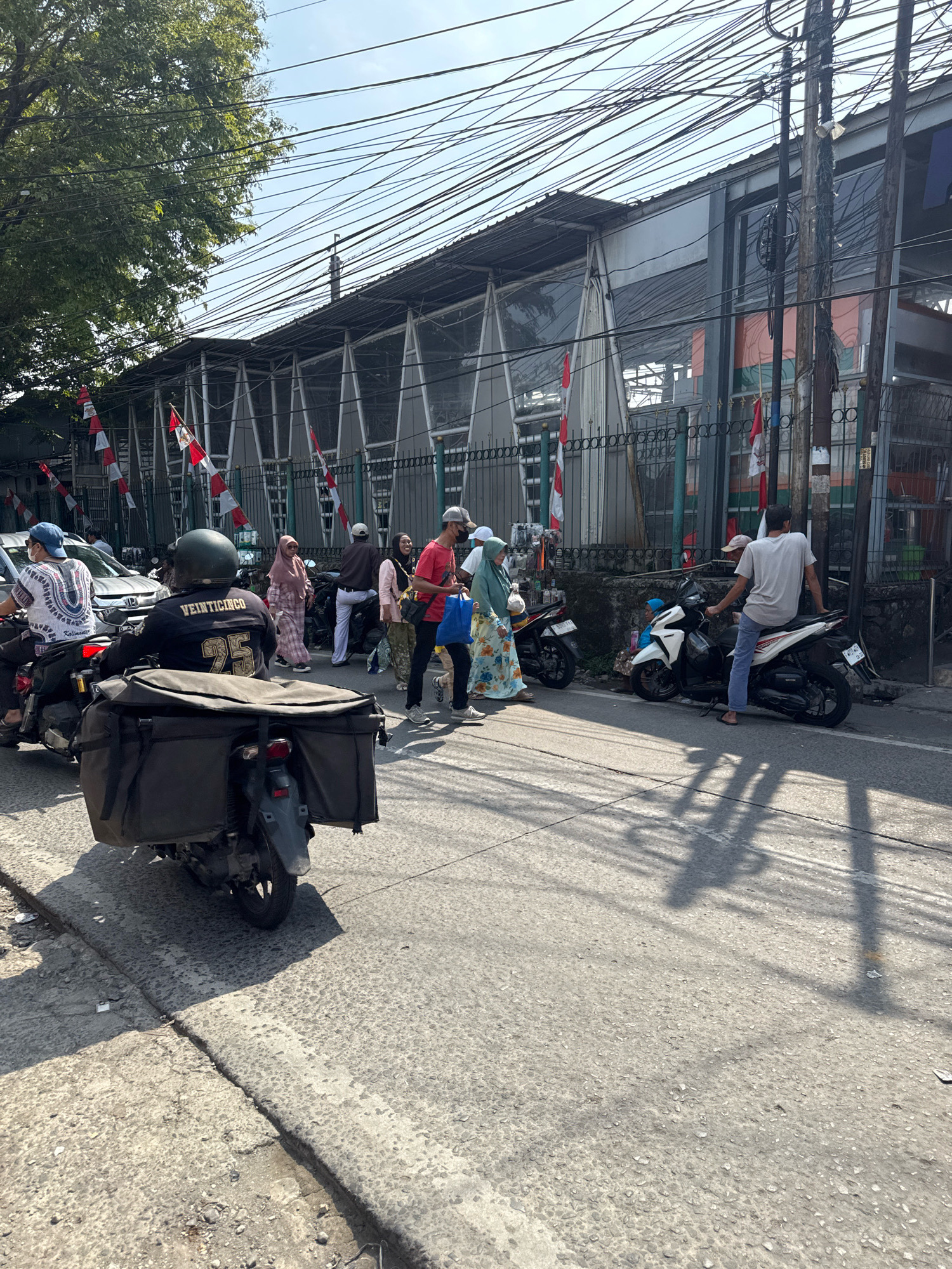 In Sukaraja, Indonesia, a street scene unfolds under a bright sky, with a complex network of overhead wires dominating the upper portion of the frame. The street itself is paved and shows signs of wear and tear, with cracks and unevenness. On the left side of the street, a person wearing a blue cap and a white patterned shirt is riding a motorcycle. Directly behind this motorcycle, another rider, wearing a dark helmet and a black jacket with the text "VEINTICINCO 25" on the back, is on a motorcycle equipped with a large, dark cargo box. Several other motorcycles are visible, some parked and others being ridden.
A group of pedestrians is crossing the street. Among them, a woman in a pink hijab and a patterned dress, another woman in a light blue hijab and a floral dress, and a man in a red t-shirt and a light-colored baseball cap are particularly noticeable. The man in the red shirt is carrying a blue bag. Another individual, a man wearing a grey t-shirt and blue jeans, is sitting on a white motorcycle on the right side of the street. Behind the people, a fence with a decorative pattern lines the sidewalk, and a building with a reflective facade and a sloping roof stands further back. Small Indonesian flags, alternating red and white, are attached to poles along the fence, suggesting a festive or nationalistic occasion. The overall impression is a bustling urban environment, typical of a market area or a busy intersection.