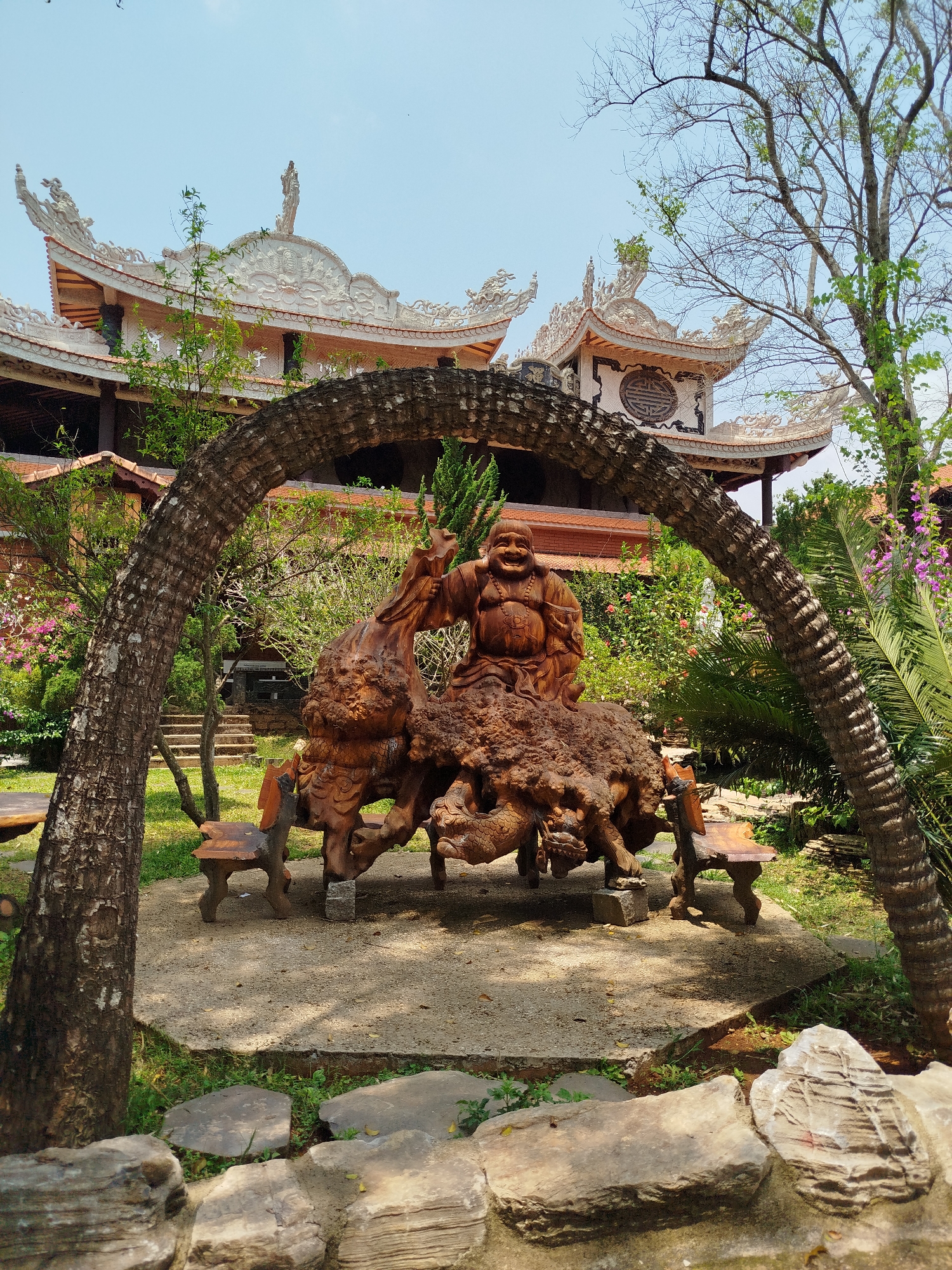 This image captures a serene garden scene in Thôn 6, Vietnam, centered around a large, intricately carved wooden sculpture. A prominent wooden statue of a jovial Laughing Buddha (Hotei), with a wide, beaming smile and making a peace sign with his right hand, is seated on a massive, gnarled wooden base that appears to be sculpted from intertwined roots or mythical creatures, possibly dragons. Several small, rustic wooden benches are arranged around the base of the sculpture.
The sculpture and benches are framed by a natural archway formed by two thick, curved tree trunks or logs in the mid-foreground. The ground directly around the sculpture is a paved circle of light-colored gravel, transitioning to grass and dirt paths. Large, flat rocks are visible in the immediate foreground.
Behind the central sculpture, a lush garden unfolds with various types of trees and flowering bushes, displaying blooms in shades of pink, purple, and red. Some trees have sparse branches with new green leaves, while others are full and verdant.
In the background, a grand, multi-tiered traditional building, characteristic of Vietnamese temple architecture, stands under a clear blue sky. Its ornate roofs are adorned with white, intricately carved decorations, including dragon-like figures and statues along the ridges and eaves. Red-tiled sections and dark wooden pillars are also visible, contributing to the building's striking appearance. One section of the building features a circular emblem with unidentifiable symbols or characters.
The scene is brightly lit by natural daylight, indicating a sunny day. The overall atmosphere is peaceful and culturally significant.