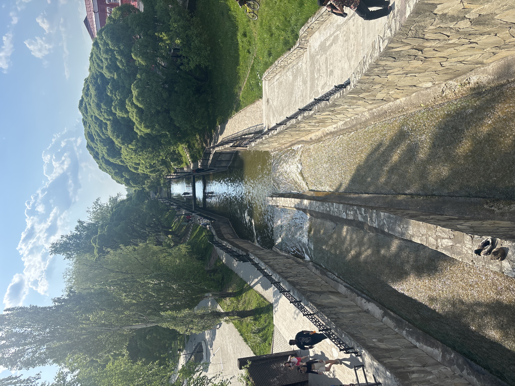 The image captures a vertical view of the Perm city embankment in Russia, specifically the part featuring a long, narrow canal with flowing water. The canal is lined with stone and concrete structures. On both sides of the canal, there are pedestrian walkways with metal railings. Trees and green vegetation are present on both sides of the canal, adding a touch of nature to the urban landscape. A group of people is visible on one of the walkways, standing near a bench. The sky is visible in the top portion of the image, with a clear blue expanse and some scattered clouds. The overall impression is a peaceful urban setting with a canal as a central feature.