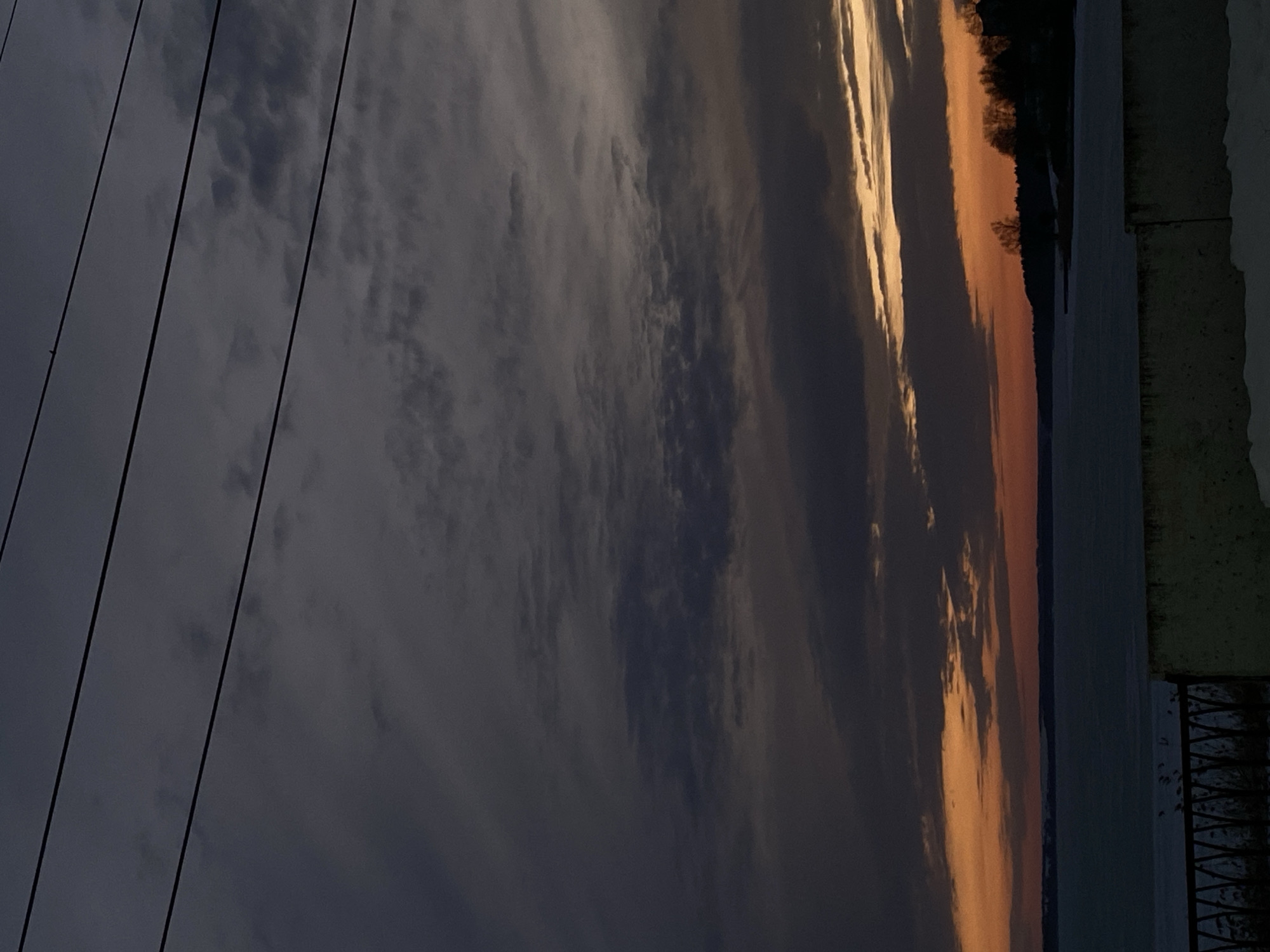 The image shows a sunset scene near the city of Votkinsk, Russia. In the foreground, power lines run diagonally from the upper left. A body of water reflects the orange and golden hues of the sunset. A treeline can be seen along the edge of the water. To the right, part of a structure or railing is visible. The sky is filled with dark, layered clouds, contrasting with the lighter colors near the horizon.