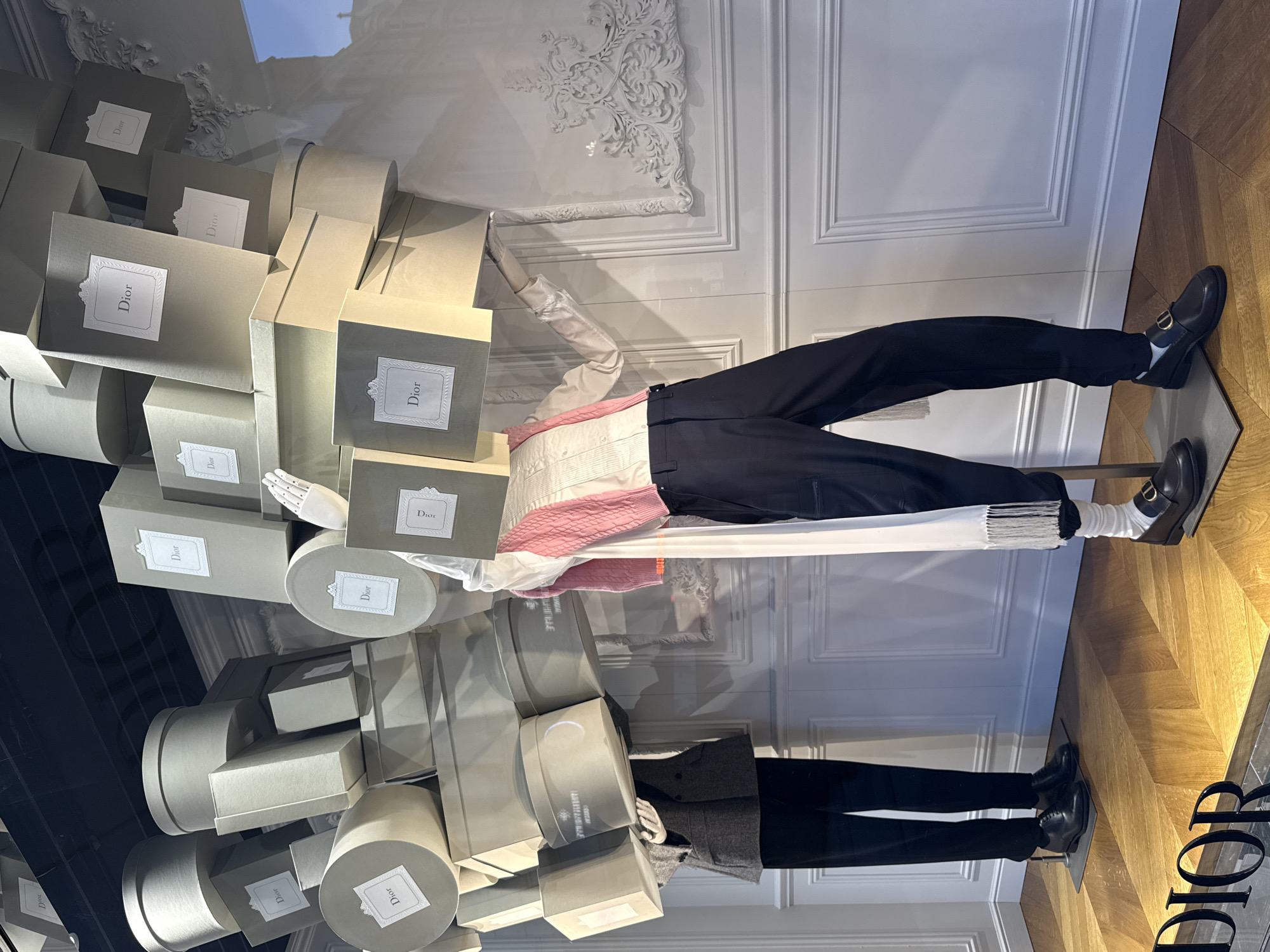 The image displays a luxury retail window display, likely from a Dior boutique on a high-end shopping street such as New Bond Street in London, United Kingdom. Two headless mannequins are positioned on a light wooden herringbone floor against a white paneled wall with decorative moldings.
The mannequin on the left wears a dark grey double-breasted jacket, black trousers, and black lace-up shoes. Its hands, covered in white gloves, appear to support a substantial, precarious stack of light grey-green gift boxes of varying circular, square, and rectangular shapes. Each visible box features a white label with "Dior" text.
The mannequin on the right wears a white long-sleeved shirt with a pleated front, a pink knitted vest, black trousers, white socks, and dark loafers with a gold-tone "CD" logo on the vamp. A white fringed scarf hangs over its left arm. Its right hand, also gloved in white, supports a similar, large stack of "Dior" branded boxes positioned above its "head."
The background wall behind the right mannequin includes an ornate, empty white frame of decorative plasterwork. Reflections on the window glass indicate an exterior architectural structure consistent with a classic urban building. A dark, dimensional "Dior" logo is partially visible at the bottom of the display window.