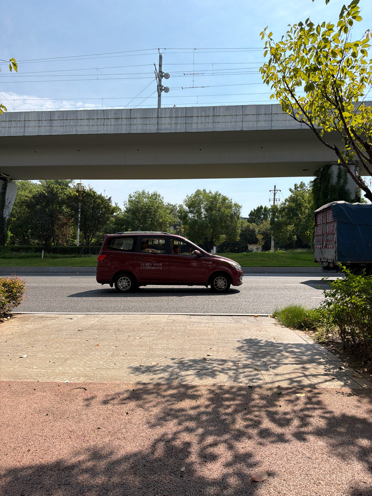 An outdoor, eye-level, medium shot captures a red minivan driving on a road under an overpass. The sky is clear and blue. Overhead, train tracks and power lines are visible, supported by poles. The overpass, made of concrete, casts a shadow on the road and the parked minivan. The minivan is in the left lane of a two-lane road, and a large truck with a blue tarp is in the right lane, moving in the same direction. To the left of the road, a sidewalk is visible, with greenery and a bush on the right side. The ground of the sidewalk is paved with two different textures, one light gray and one reddish-brown, and is dappled with the shadow of tree leaves. The minivan has its windows down, and two individuals are visible inside. The truck's side has Chinese characters on it, indicating it is likely in China. The presence of Chinese characters on the truck and the overall aesthetic of the infrastructure suggest the location is within Jiangjin, China.