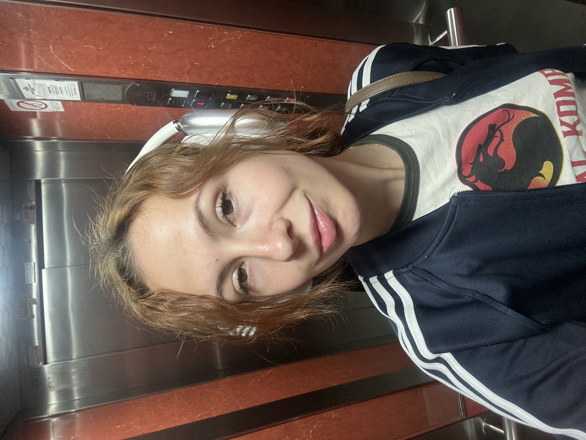 A young woman is taking a selfie in an elevator with a wood-grain panel. She has shoulder-length, reddish-brown hair. She is wearing white wireless headphones, a white T-shirt with a "Mortal Kombat" logo on the front, and a dark blue Adidas track jacket. The elevator has stainless steel walls on either side of the wood-grain panel. Elevator buttons are visible above the panel. The location is likely a residential or commercial building in Novi Sad, Serbia.