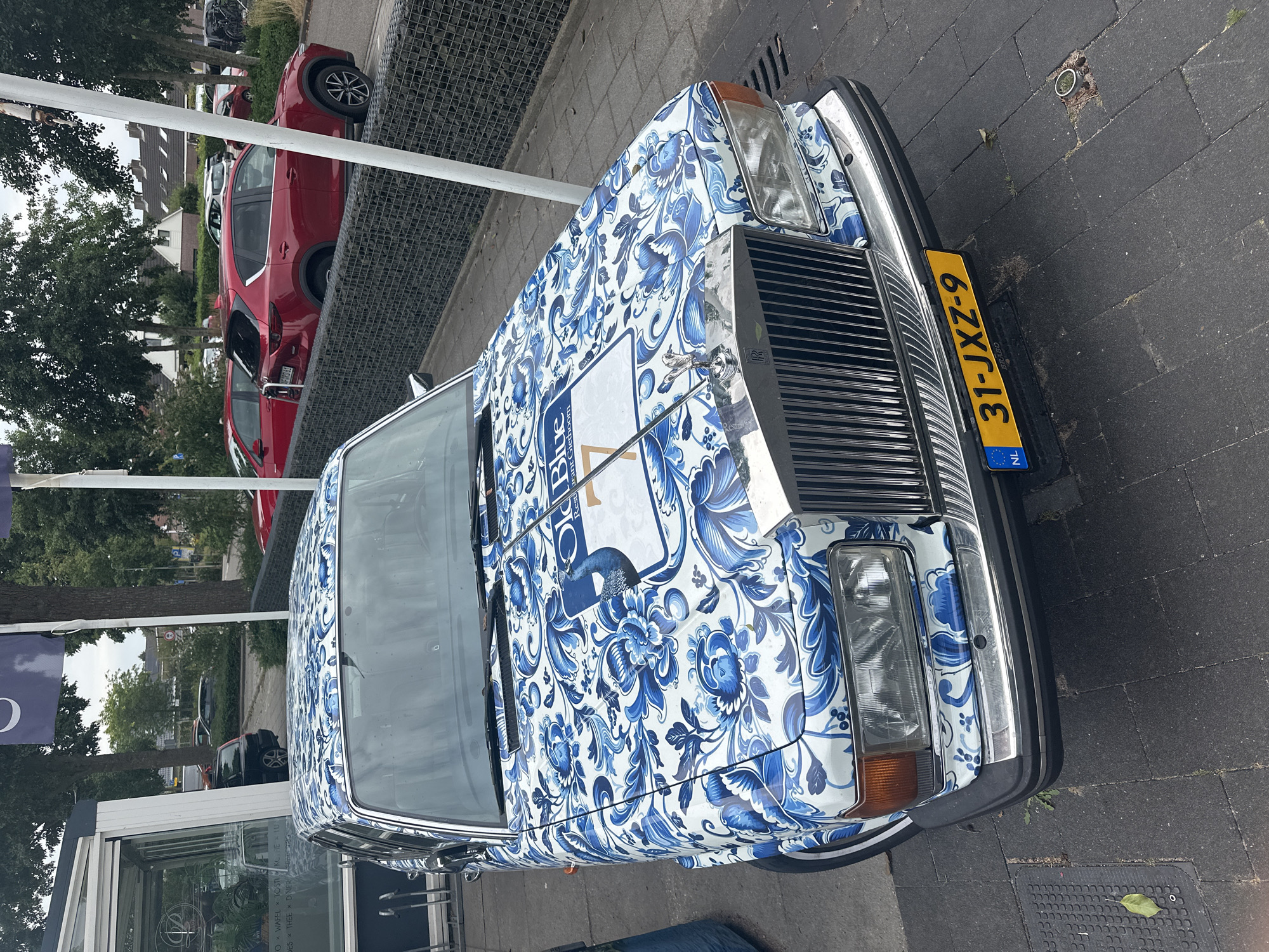 The image shows the front and hood of a Rolls-Royce car parked on a paved walkway. The car is covered in a blue and white floral Delft blue pattern. A white sign with blue text "Old Blue Restaurant Giethoorn" and a blue logo featuring a bird is visible on the hood. A license plate with the number "31-JXZ-9" is attached to the front bumper. In the background, there are trees, a building with a glass facade, and other parked cars, including a red Mazda SUV. The setting appears to be a public area in Giethoorn, Netherlands, likely near the restaurant mentioned on the sign. The lighting suggests it is daytime, with overcast skies.