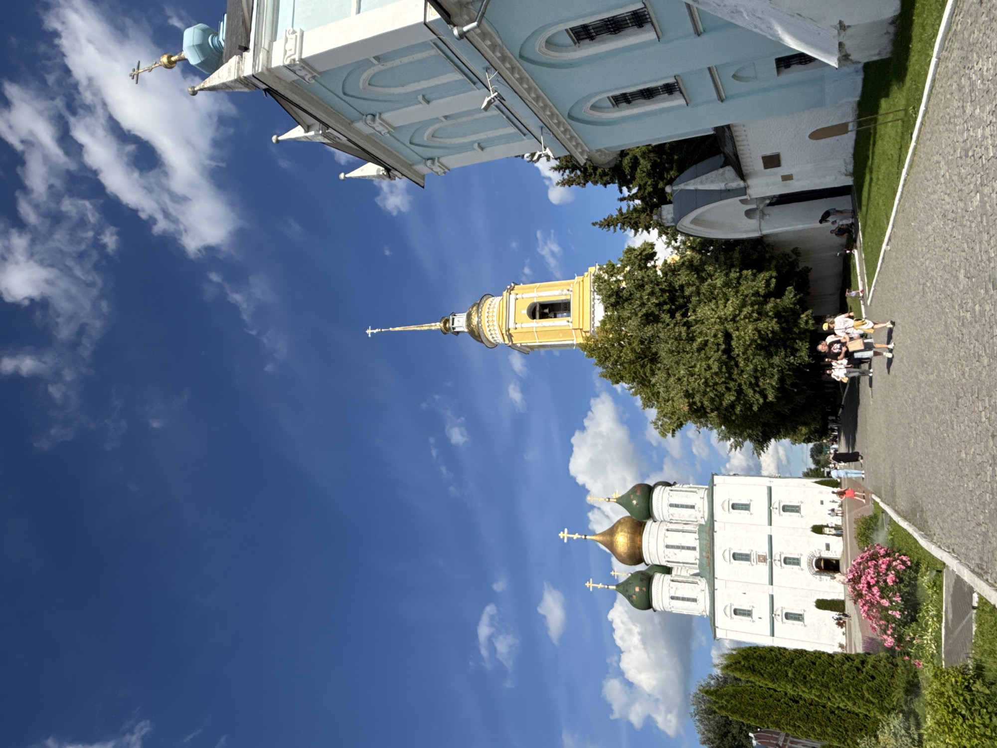 The image shows a sunny day with a blue sky filled with scattered white clouds. In the foreground, a paved walkway leads towards several buildings. On the left, a large white church with green and gold domes and crosses is visible. A vibrant pink flower bush is in front of it. To the right of the white church, a tall yellow bell tower with a gold spire and cross stands prominently. Further to the right, a light blue and white building with arched windows and decorative white trim is partially in view. Several people are walking along the path, some dressed in casual clothing, appearing to be tourists. The environment suggests a historical or religious site, likely a monastery or cathedral complex.