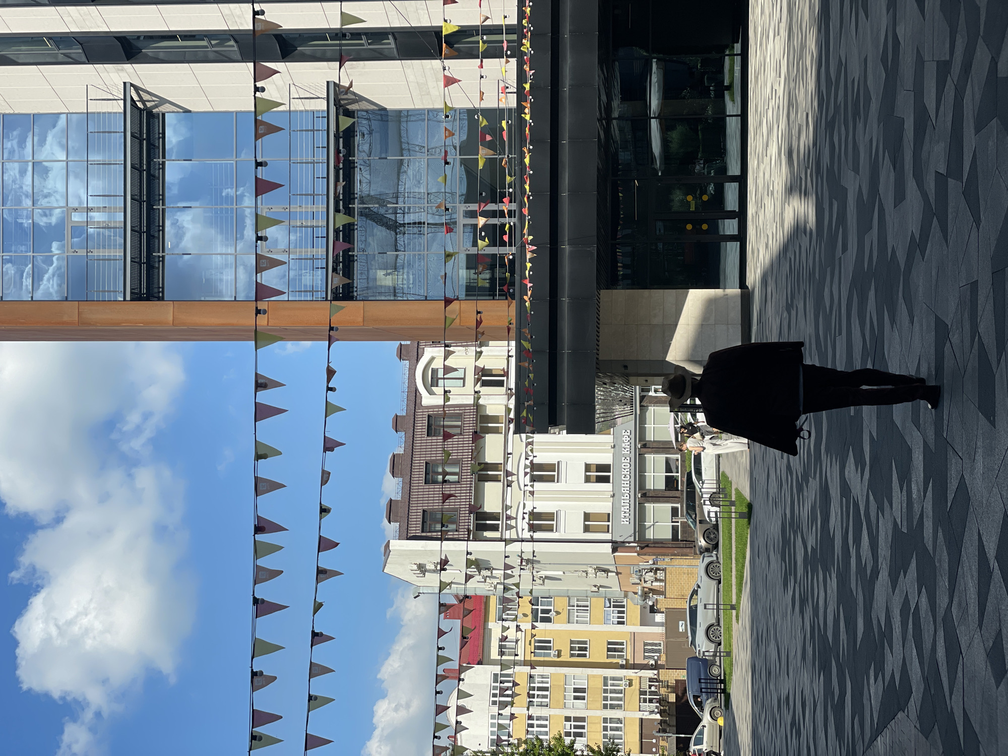 In Kazan, Russia, a person wearing a hat and a dark jacket walks away from the camera on a patterned pavement. They are in a plaza in front of a modern building with large glass windows reflecting the sky and clouds. Above the plaza, colorful triangular flags are strung across wires. To the left of the modern building, there are several older buildings with pitched roofs and numerous windows. One of these buildings features a sign that reads "ИТАЛЬЯНСКОЕ КАФЕ" (Italian Cafe). Cars are parked in the background, and a few people are visible near the cafe entrance. The sky is blue with fluffy white clouds.