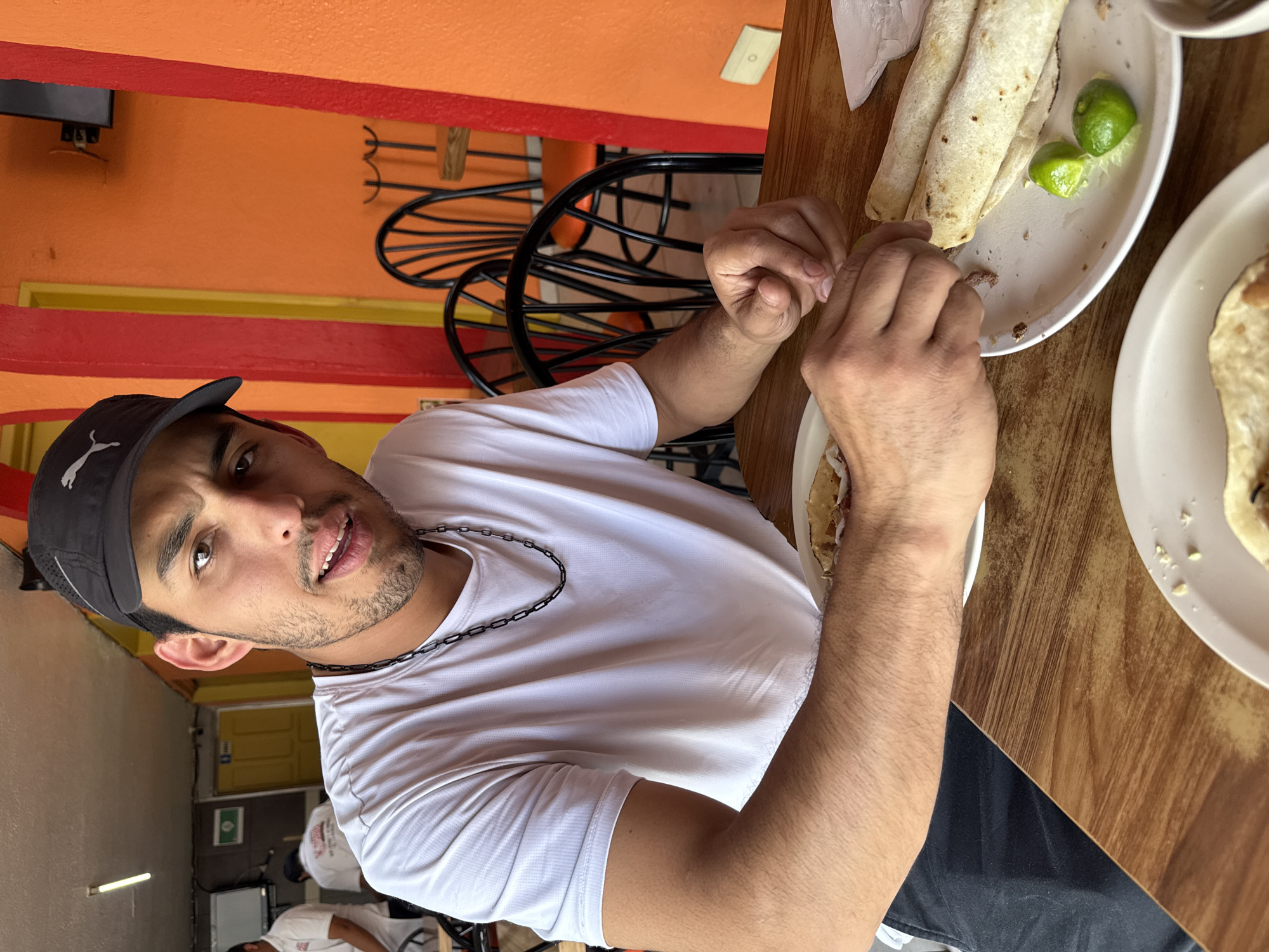 A young adult male, with short dark hair and a beard, is seated at a light brown wooden table inside a restaurant in Puebla, Mexico. He is wearing a dark grey baseball cap with a white Puma logo, a white short-sleeved t-shirt, and a black link chain necklace. He looks directly at the camera with his mouth slightly open. His left hand is positioned over a white ceramic plate containing three rolled tortillas and two green lime wedges, one of which he is pressing. Another white ceramic plate with similar food is partially visible in the lower-left foreground. A crumpled white paper napkin is on the table to the right. The background features a vibrant orange wall with vertical red columns. Two black metal chairs with curved backrests are visible, one having an orange seat cushion. A yellow door is in the left background, with a green rectangular sign above it. Two other individuals in white shirts are partially visible and out of focus in the deeper background. The ceiling is white, with an exposed fluorescent light fixture. A light-colored electrical wall plate and a dark wall-mounted object are visible on the orange wall.